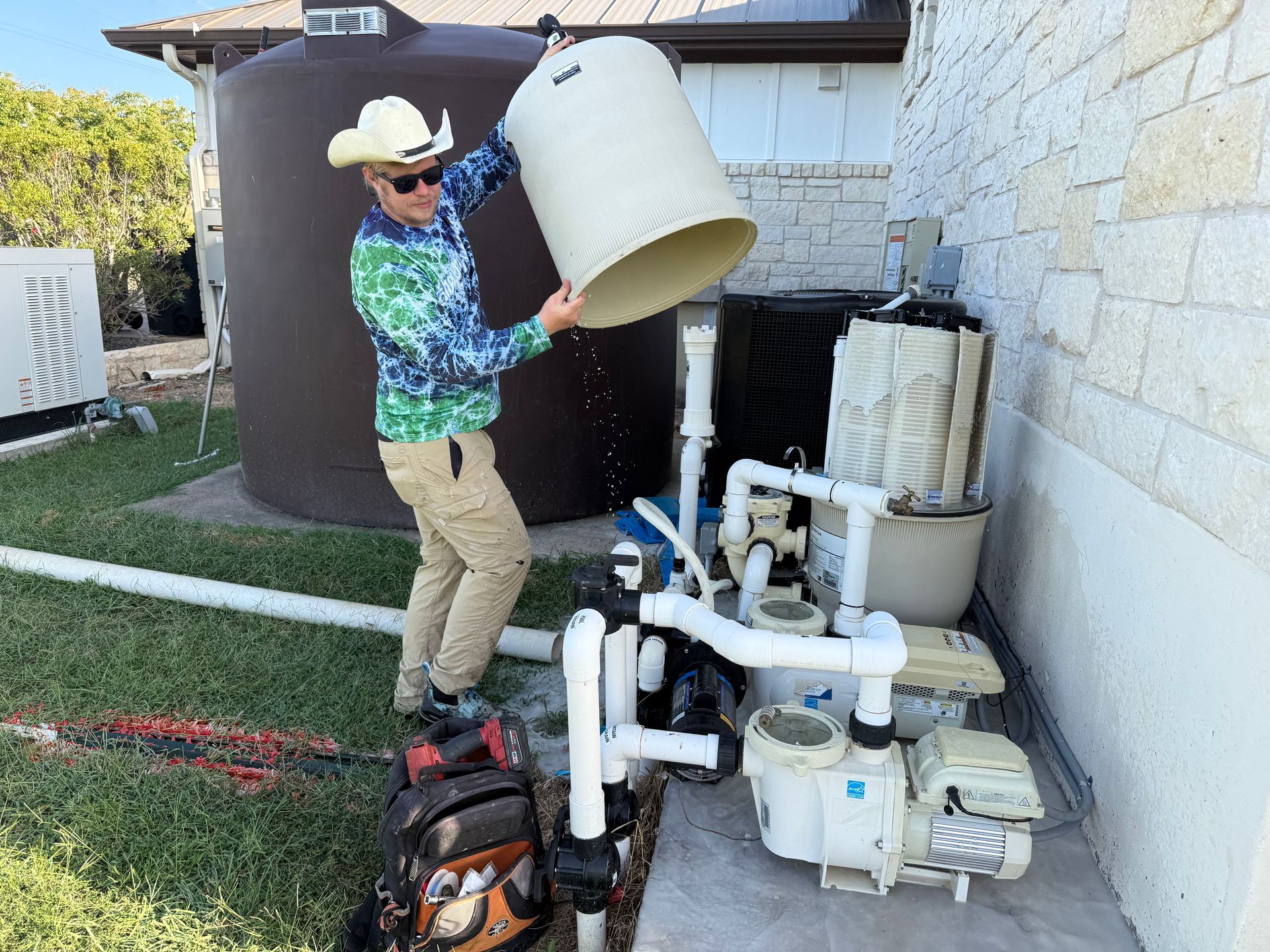 Man in cowboy hat empties a container near pool equipment outside. Dark water tank in background.