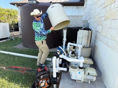 Man in cowboy hat empties a container near pool equipment outside. Dark water tank in background.