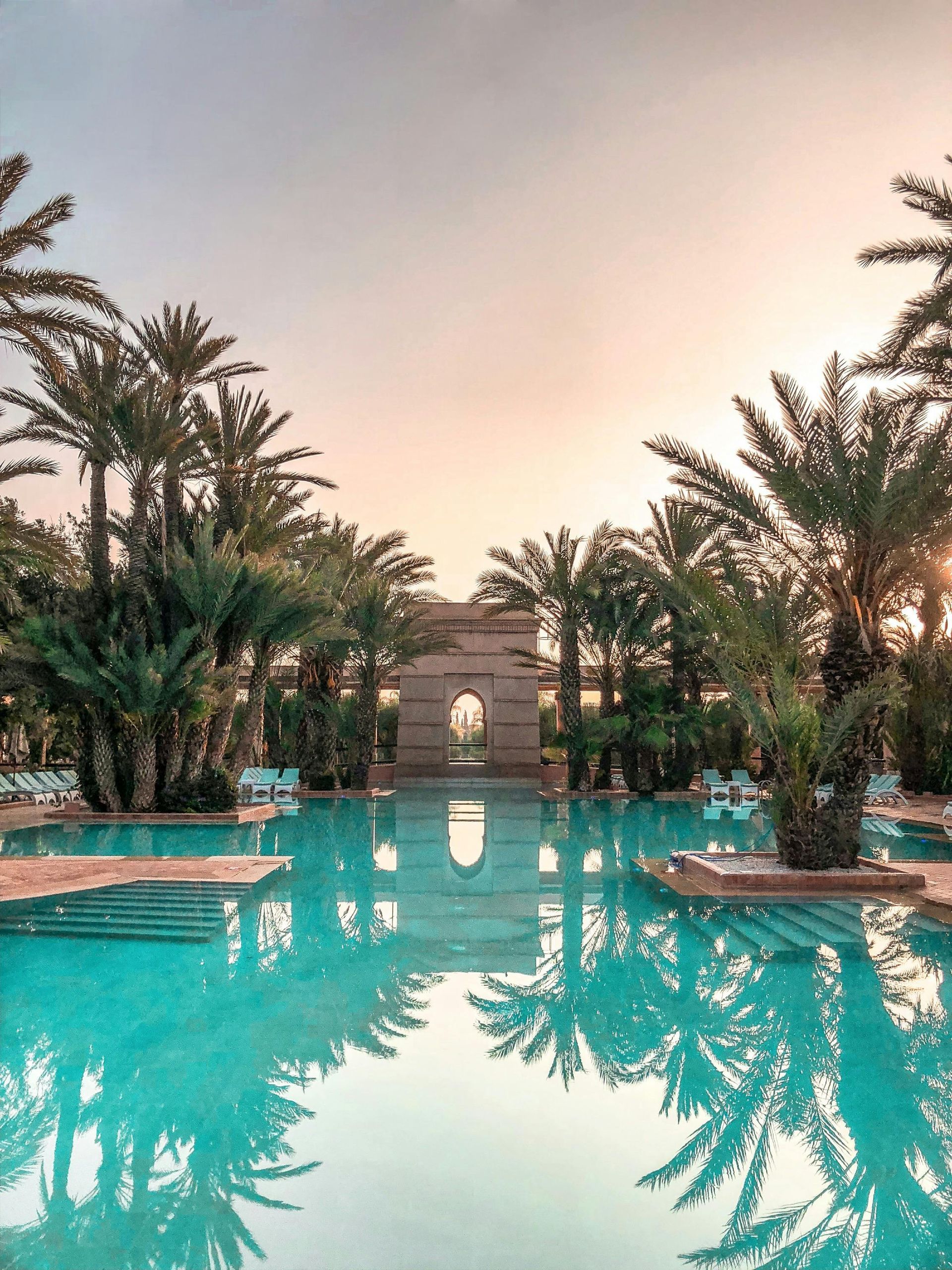 Pool with palm trees, reflecting the sky at sunset. Stone archway in the background.