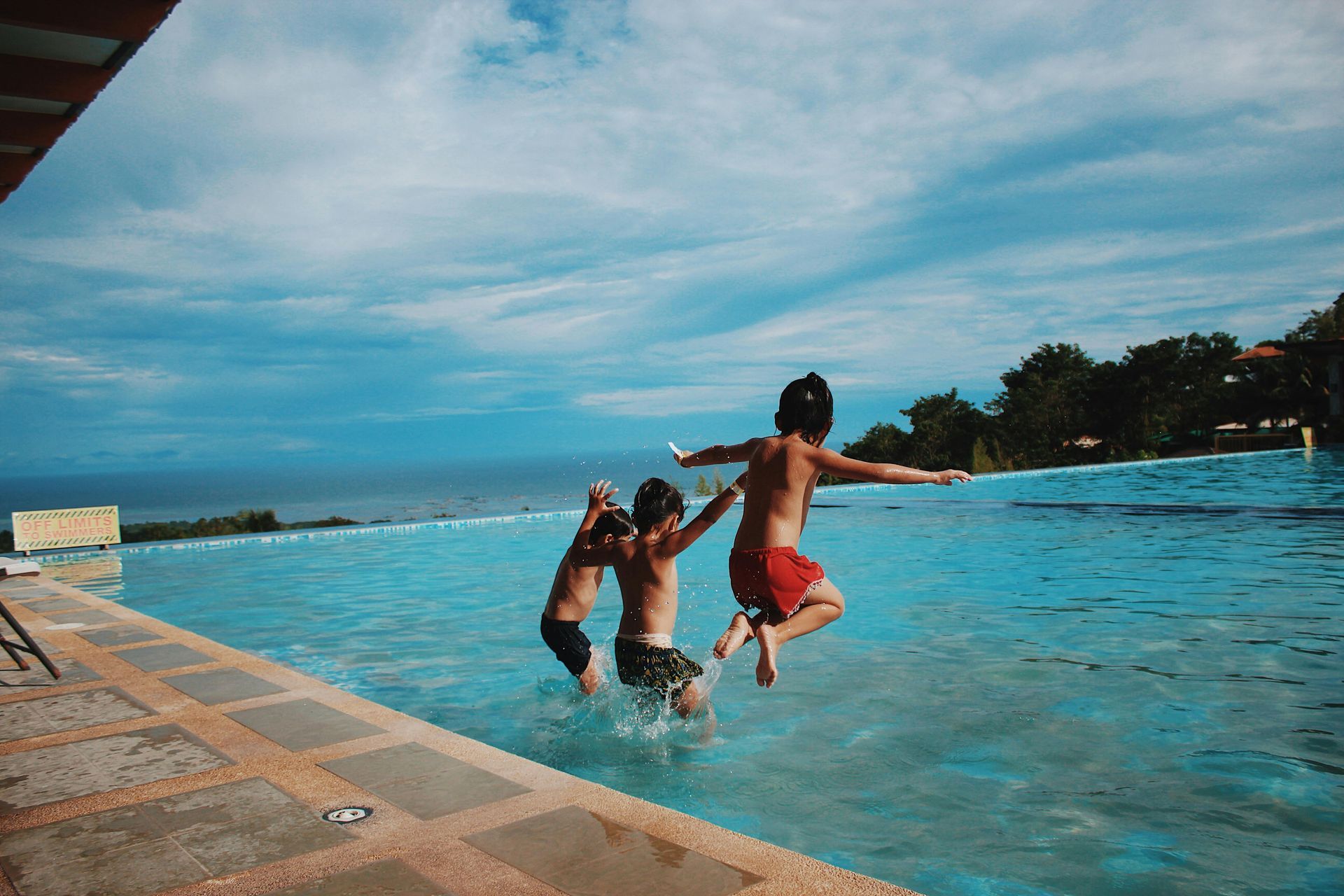 Three kids jumping into a pool on a sunny day with ocean view.