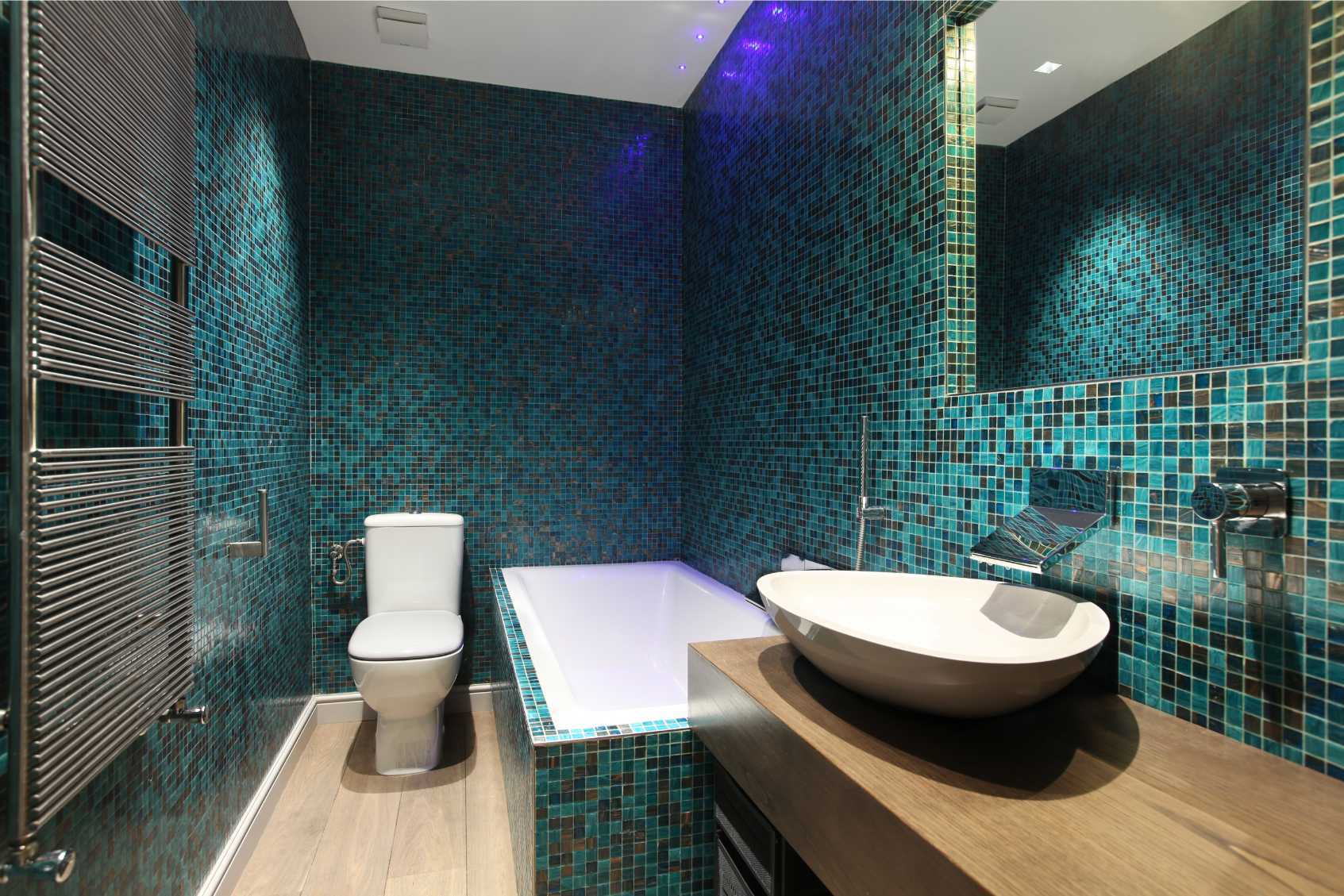 Bathroom with blue and black glass mosaic tiles. There is a bath, a toilet, a sink, a soap dispenser, a towel rail and a mirror.