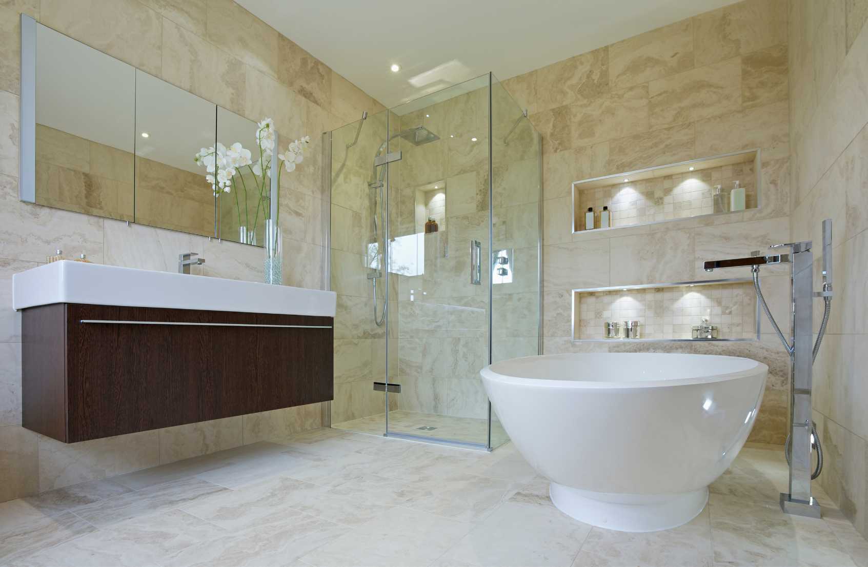 Small round bath, large glass enclosure shower in the background, a large mirror is visible above a large sink. The tiles are all made out of ceramic, finished in a cream colour.