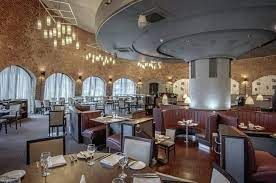Restaurant with brown seating boards, grey and stained wooden chairs, chandeliers, and laid-up tables.