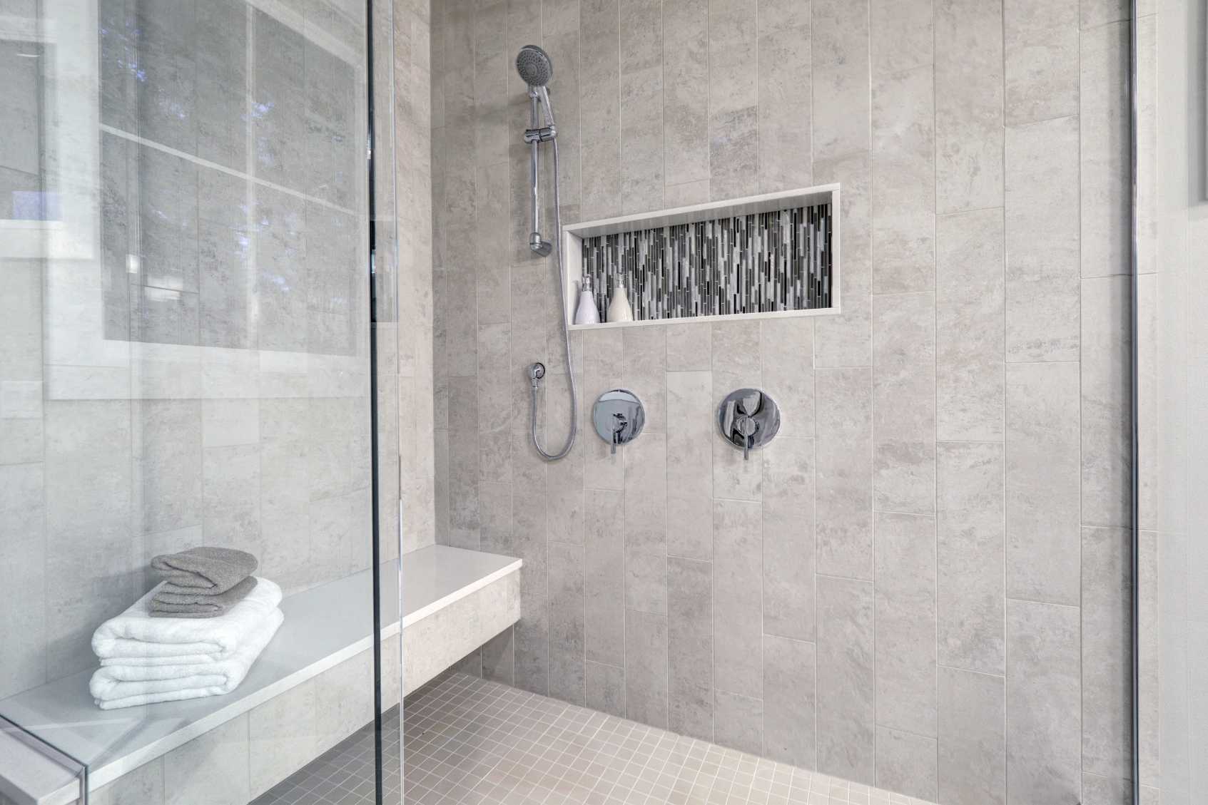 Shower with a built-in seat which has towels on it. The tiles are white and modern, the shower head is small and attached to the wall. The panel is made out of glass.