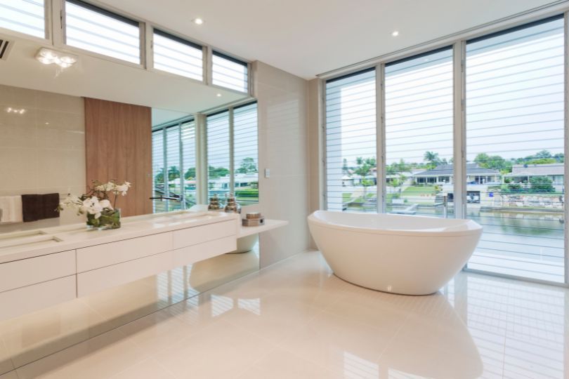 A large bathroom with a white bath, a white double sink vanities, white floor, and large windows overlooking a river.