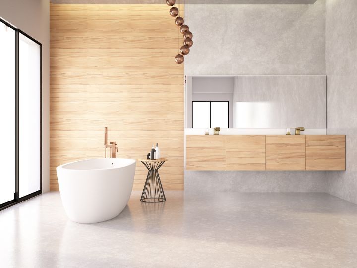A white, free-standing bath, with toiletries sat on a small table next to it. There is a large mirror in the background, the floor is grey, as well as some of the walls, the rest of the walls are made out of oak and are bright.