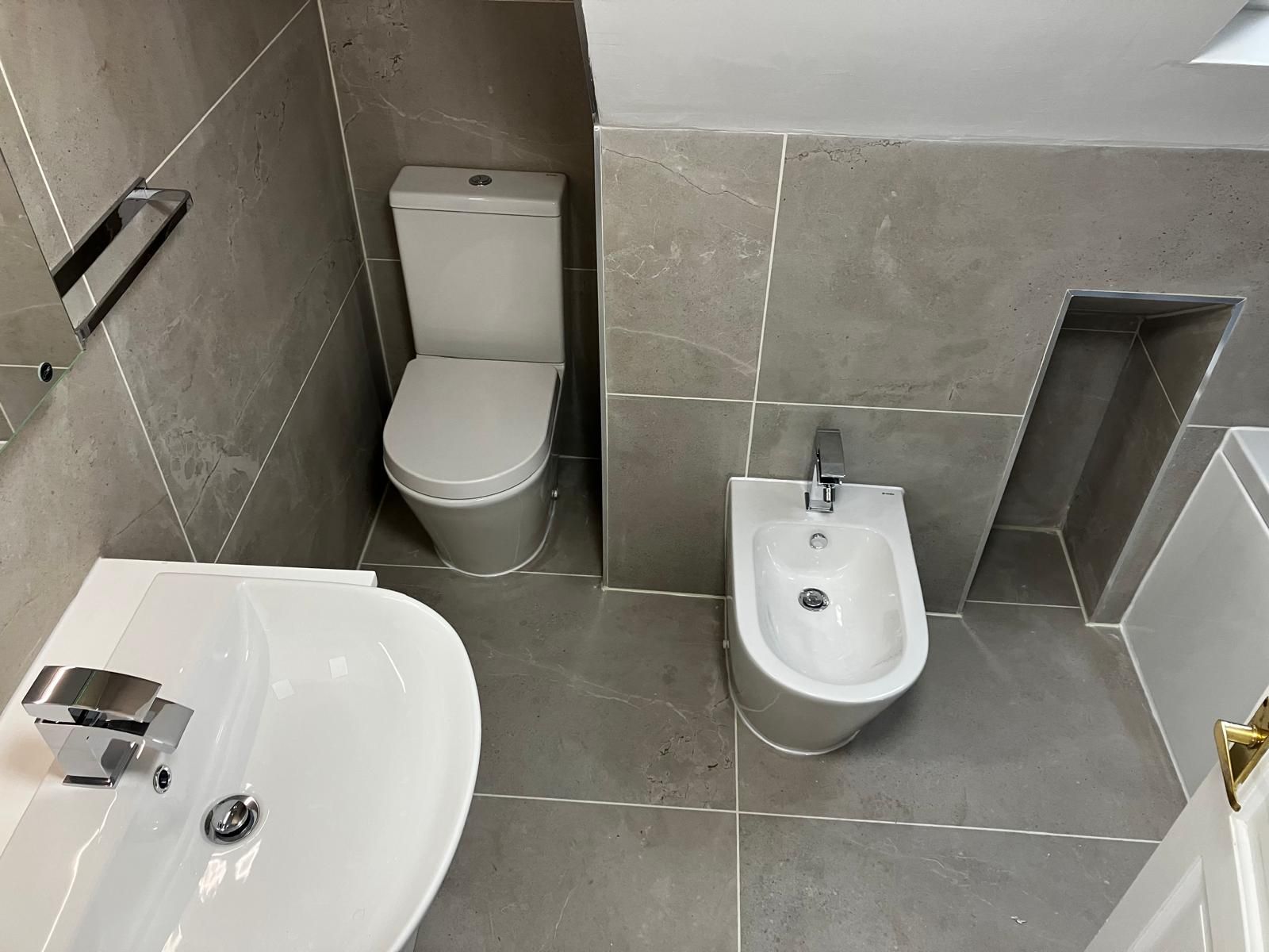A bathroom with a toilet, bidet, and sink; gray tiled walls and floor.