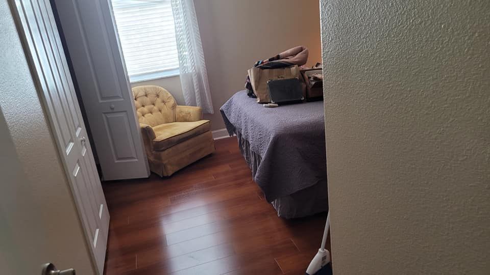 Bedroom interior with a bed, chair, and closed door. Dark wood floors, a window with blinds.