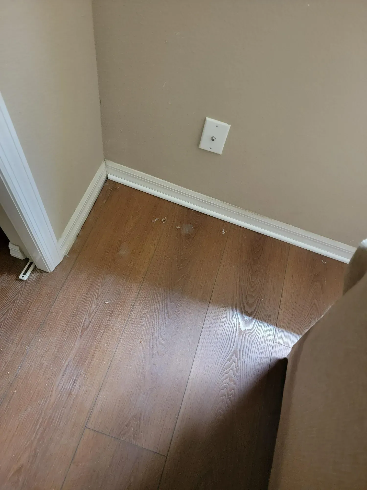 Corner of a room with brown wood-look flooring, beige walls, white trim, and a white electrical outlet.