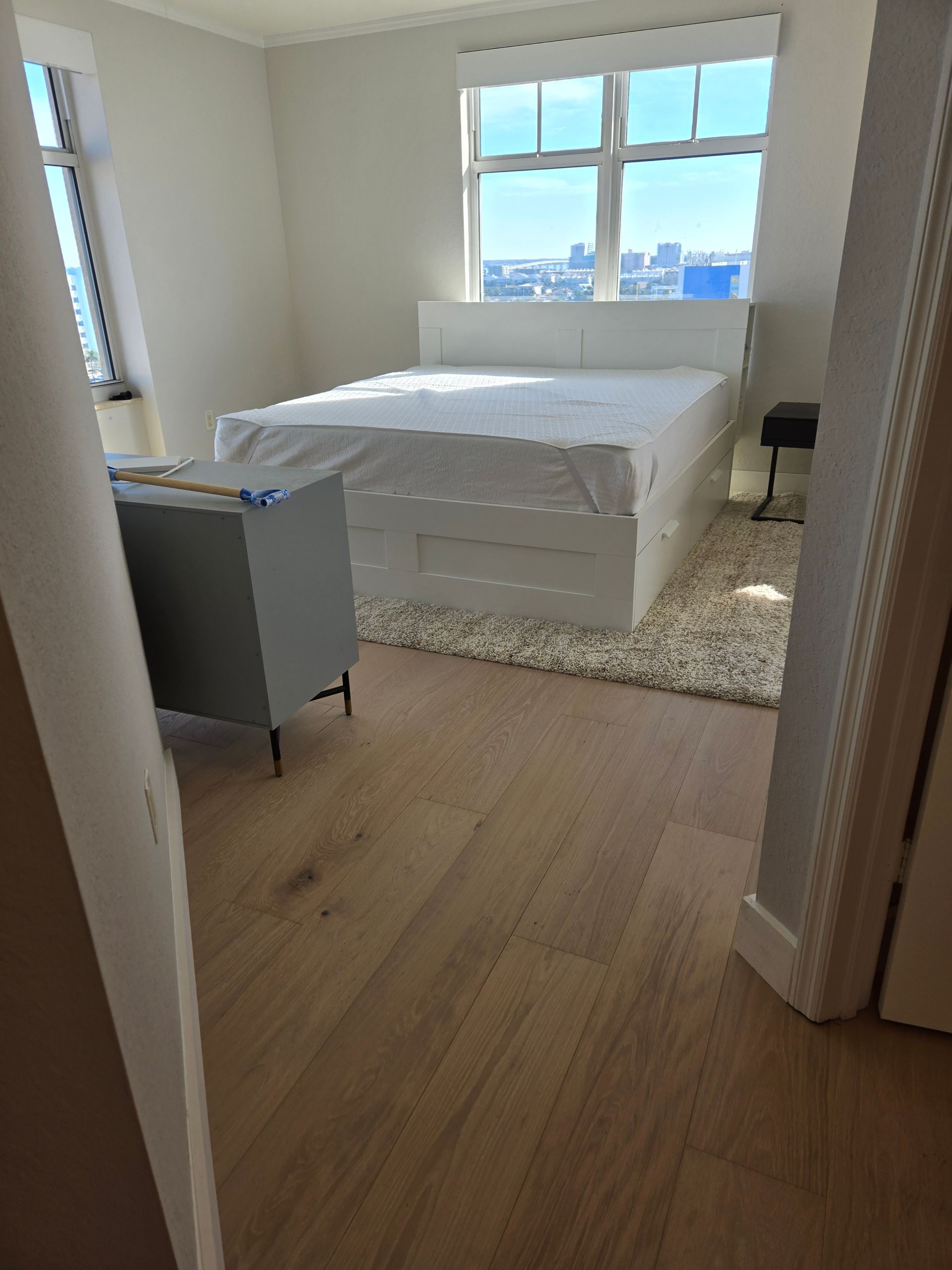 Bright bedroom with white bed, wood floor, and large window overlooking a city skyline
