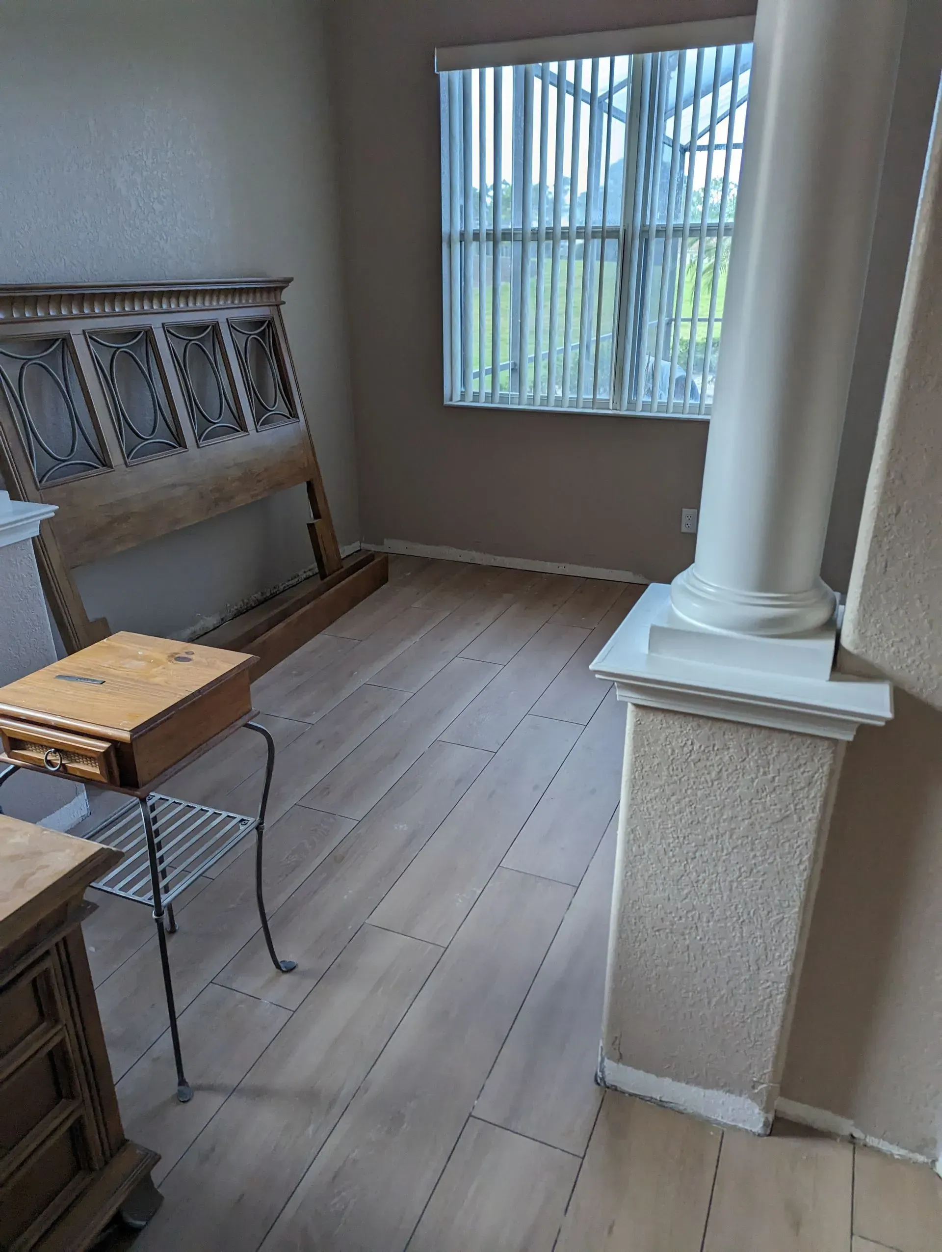 A room with a wooden headboard, small table, and a window with blinds. Light wood-look flooring.