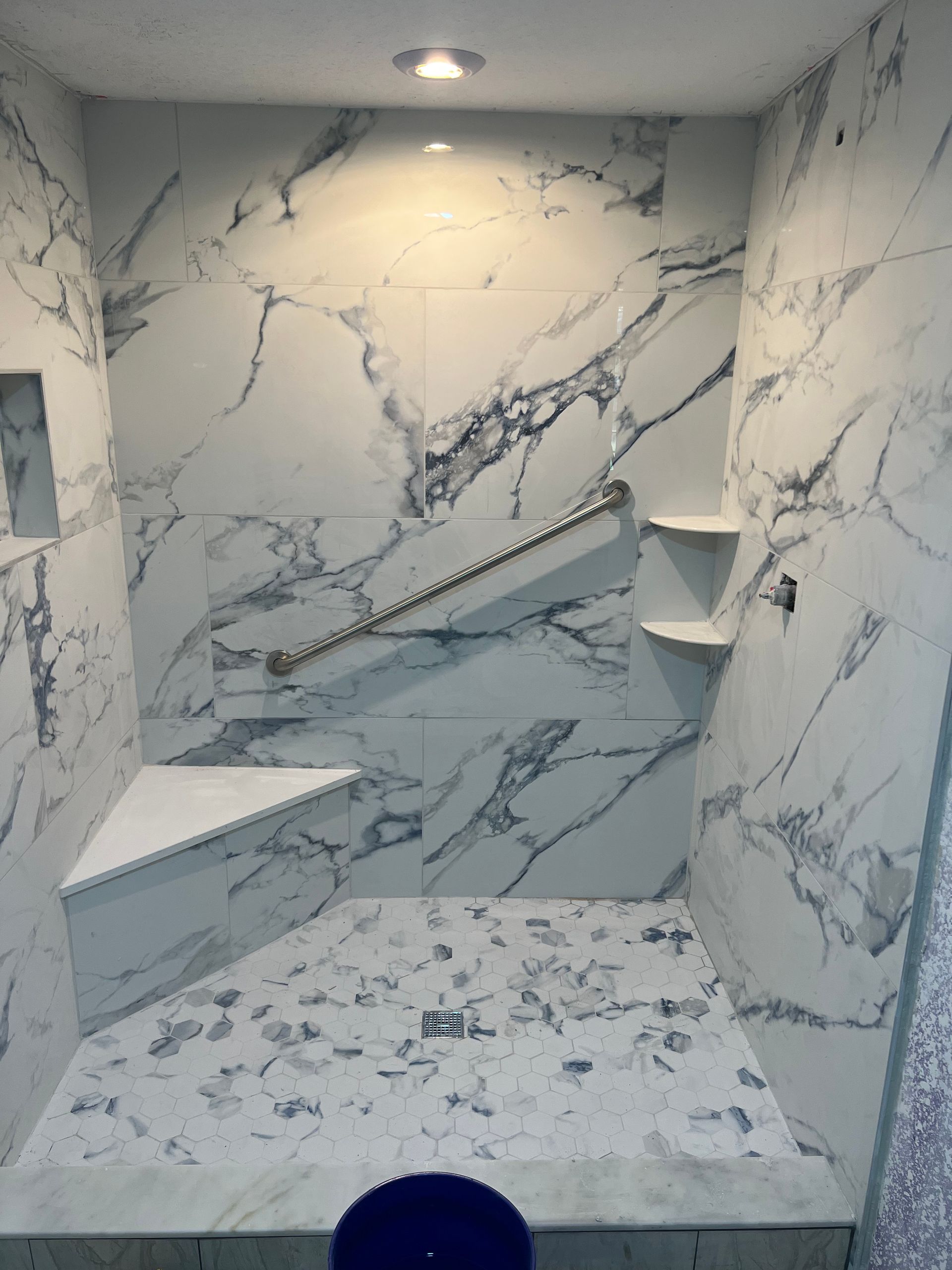 Small marble-tiled shower stall with built-in corner shelves and overhead light.