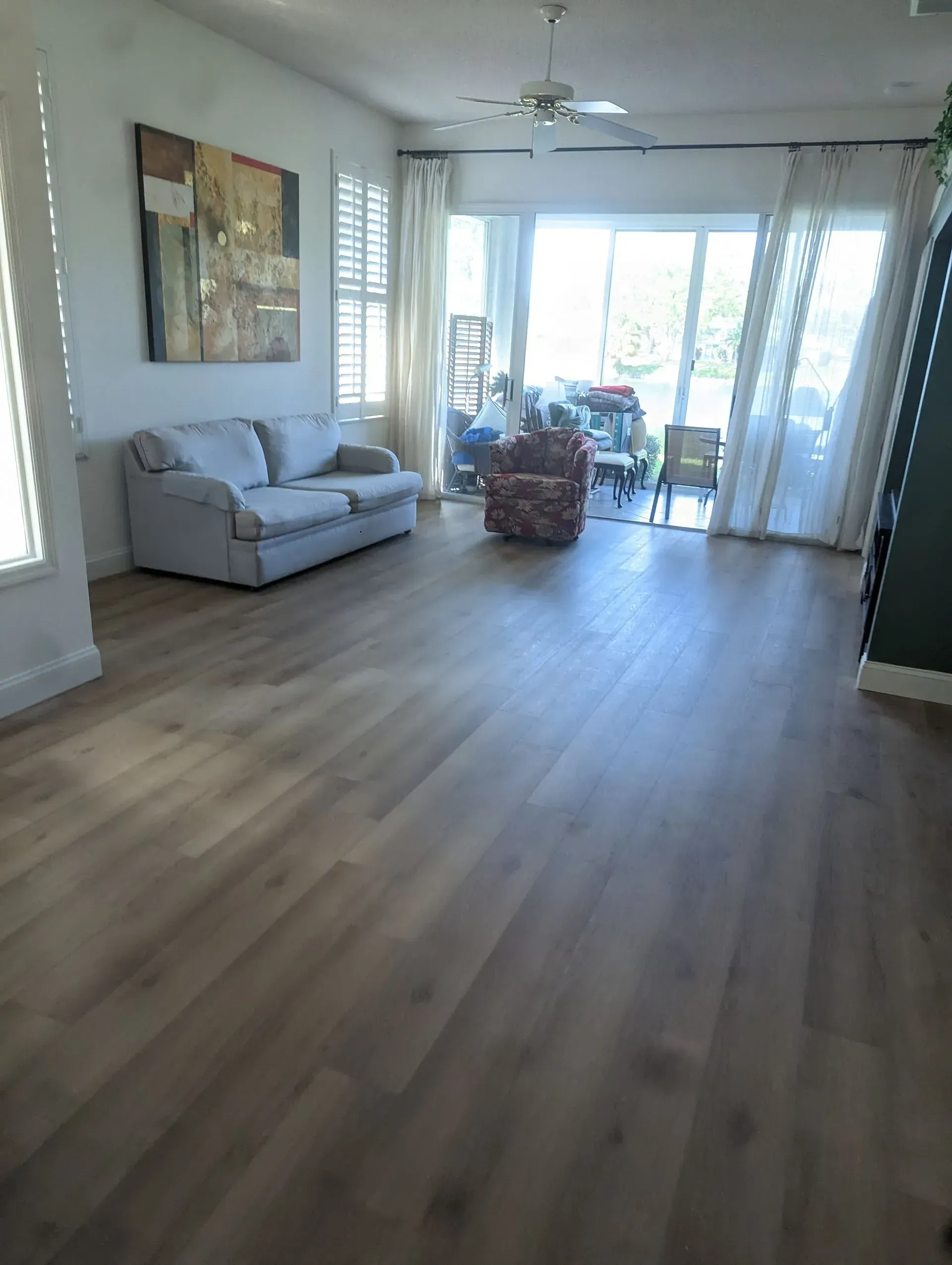 Living room with light wood floors, sofa, large windows, artwork, and a ceiling fan.