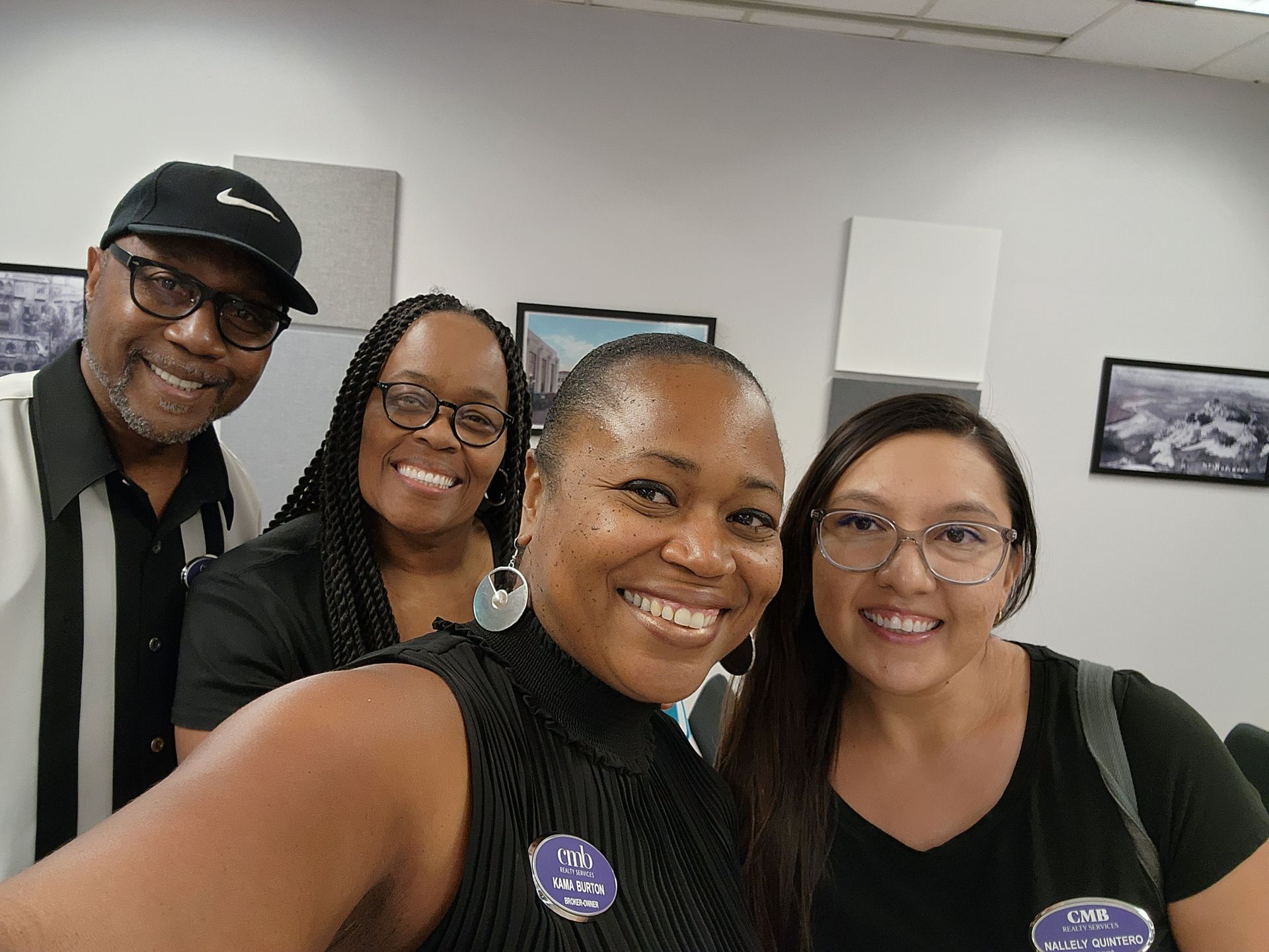 Four smiling colleagues in an office setting. From left to right: an older man wearing a Nike cap and glasses, a Black woman with braids and glasses, a Black woman with short hair wearing a name tag, and an Mexican woman with long hair and glasses. All are dressed in dark clothing. The background shows white walls with framed photographs.