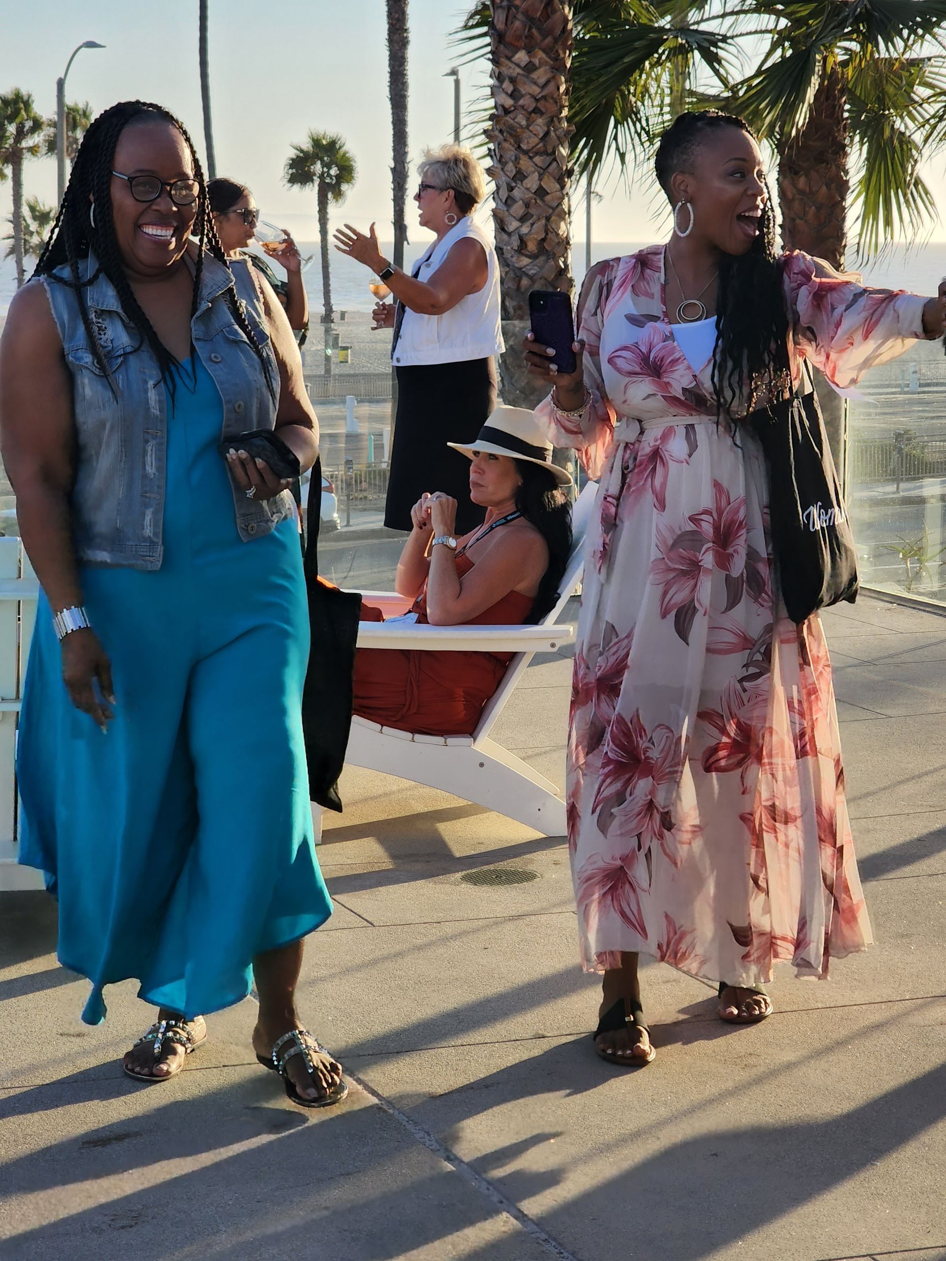 A group of women enjoying a beach boardwalk. In the foreground, two Black women in colorful dresses - one blue, one floral - are smiling and walking. Behind them, other women are visible: one sitting in a white chair wearing a hat, another standing and gesturing. Palm trees and lampposts line the walkway, with the ocean visible in the background, suggesting a warm, coastal setting.