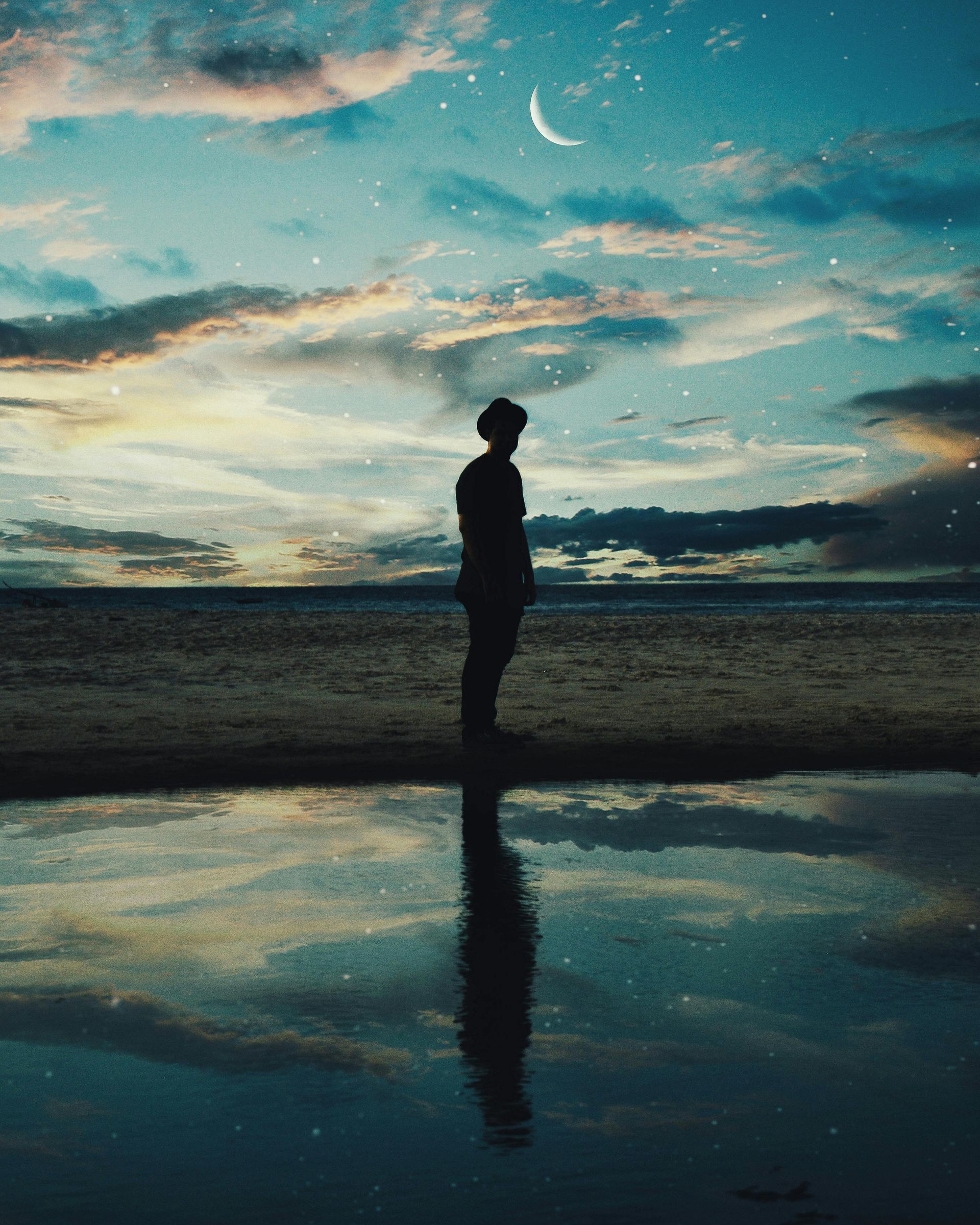 A silhouette of a person standing on a beach at sunset with a crescent moon in the sky.