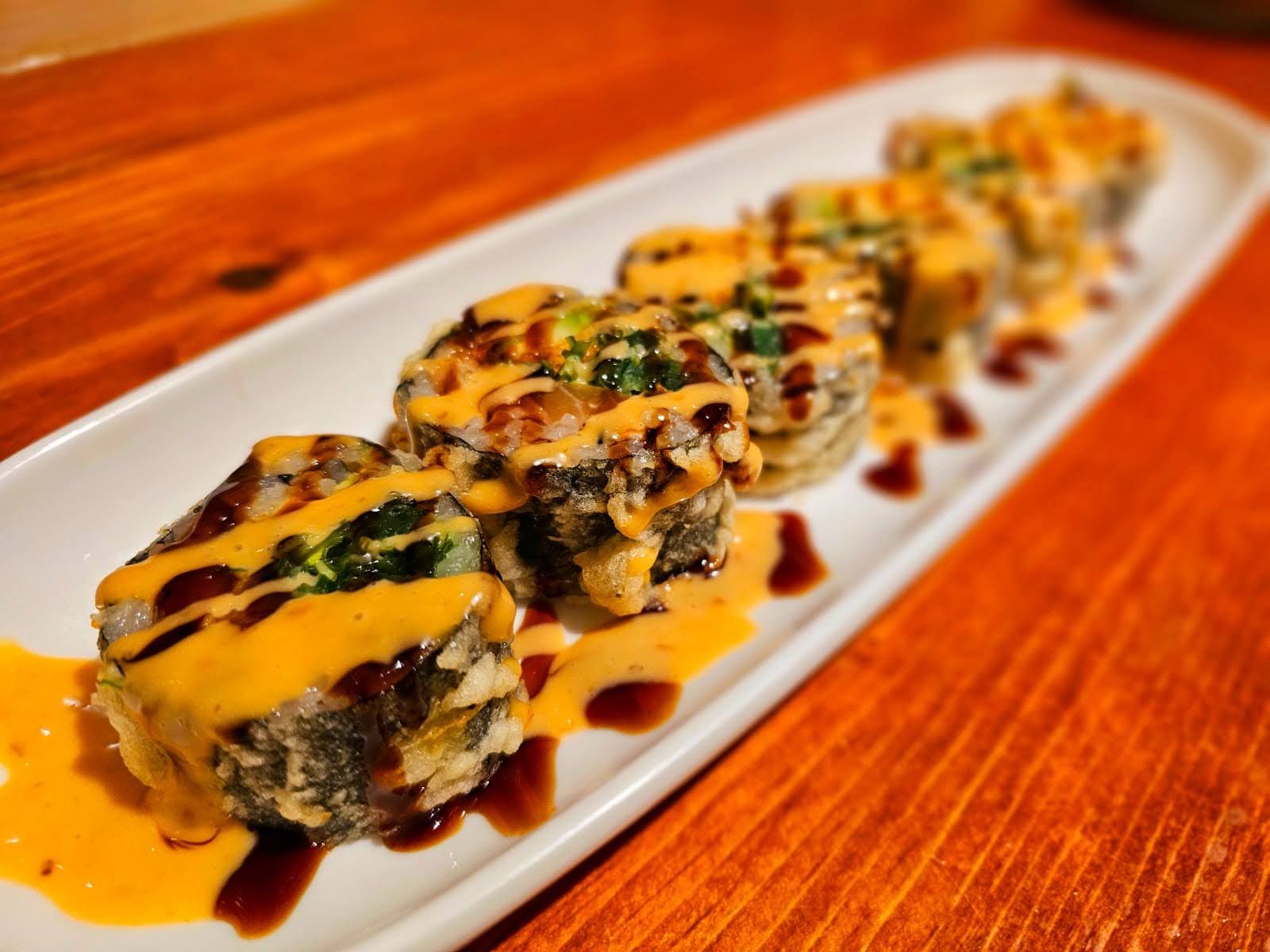 Plated sushi rolls drizzled with orange sauce on a white serving dish, on a wooden table