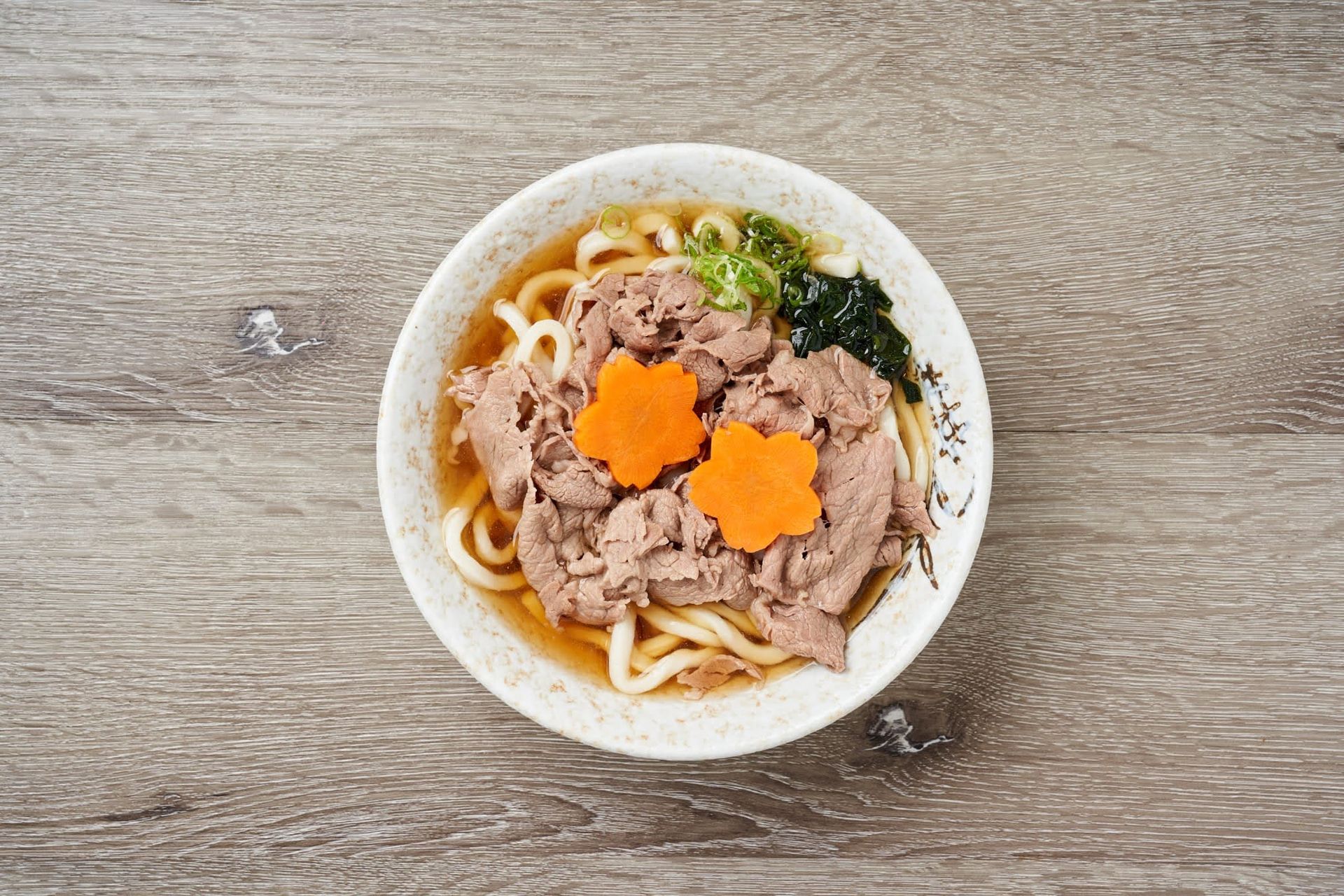 Top-down view of a bowl of noodle soup with sliced meat and orange carrot flowers on a wooden table