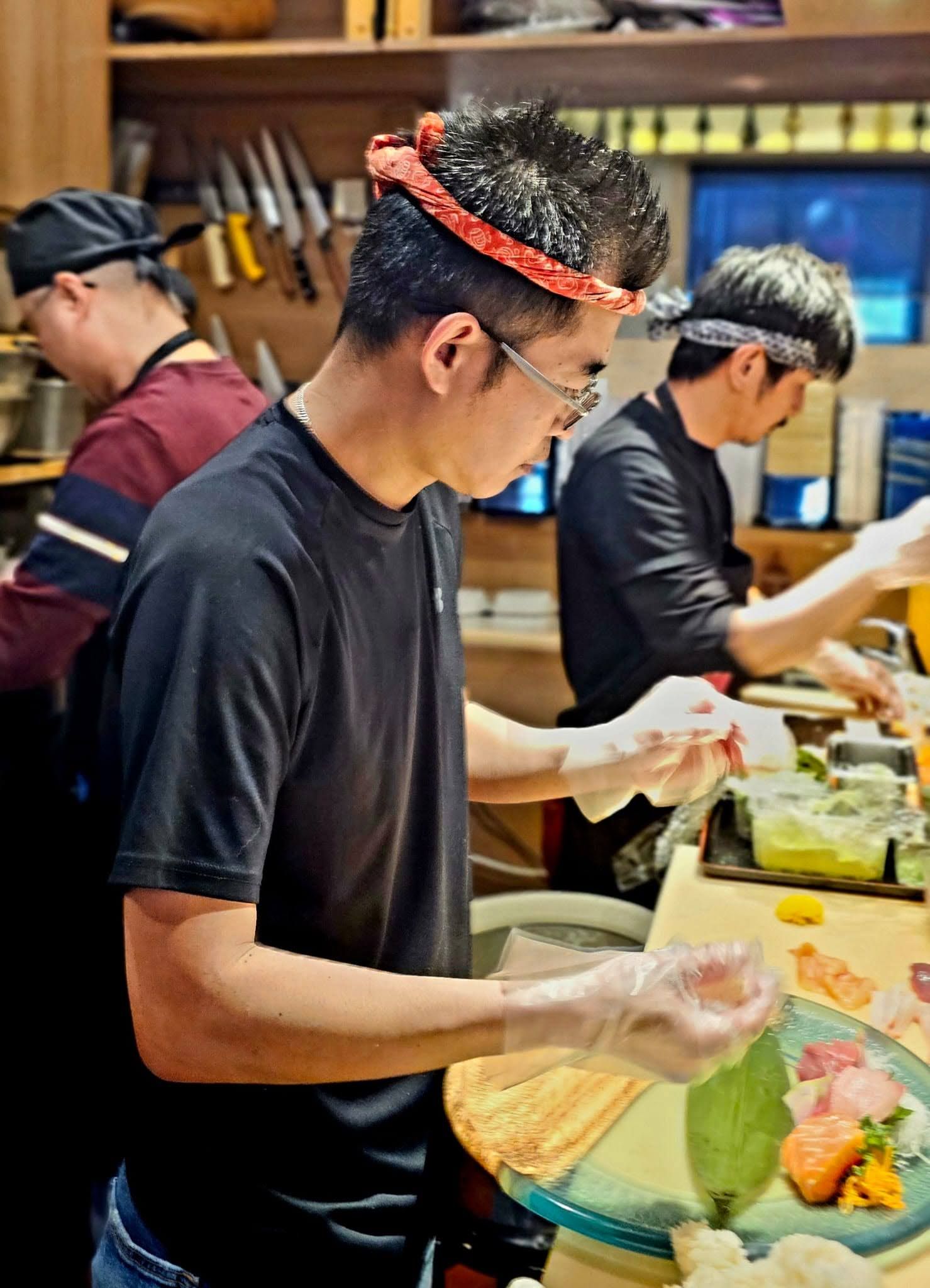 People preparing food at a buffet counter, wearing gloves and serving dishes in a restaurant.