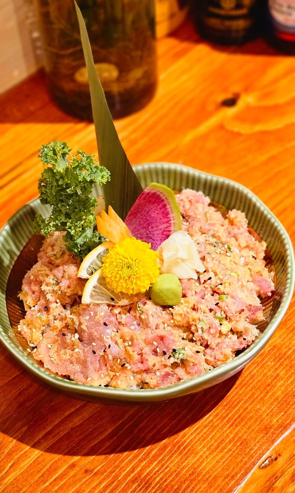 Bowl of colorful dessert with pink shaved ice, flowers, and a green leaf on a wooden table