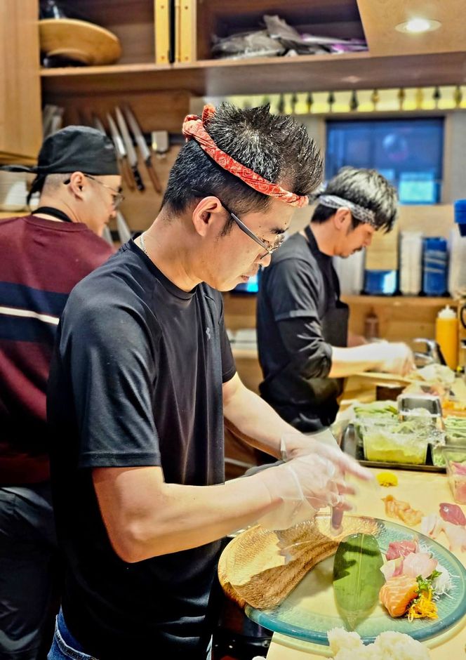 Two chefs preparing food at a counter, wearing gloves and black shirts, with colorful ingredients on a table.