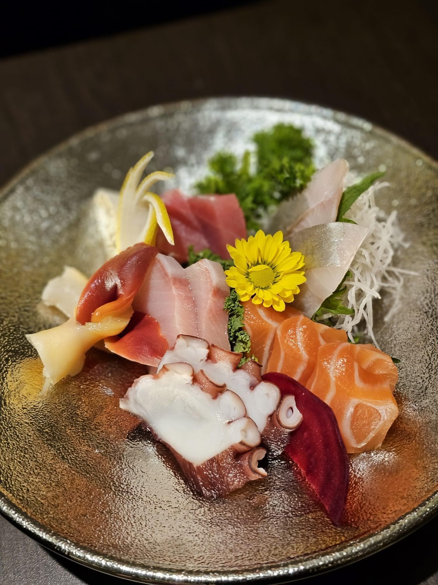 Assorted sashimi with garnishes on a ceramic plate, including salmon, tuna, octopus, and edible flowers