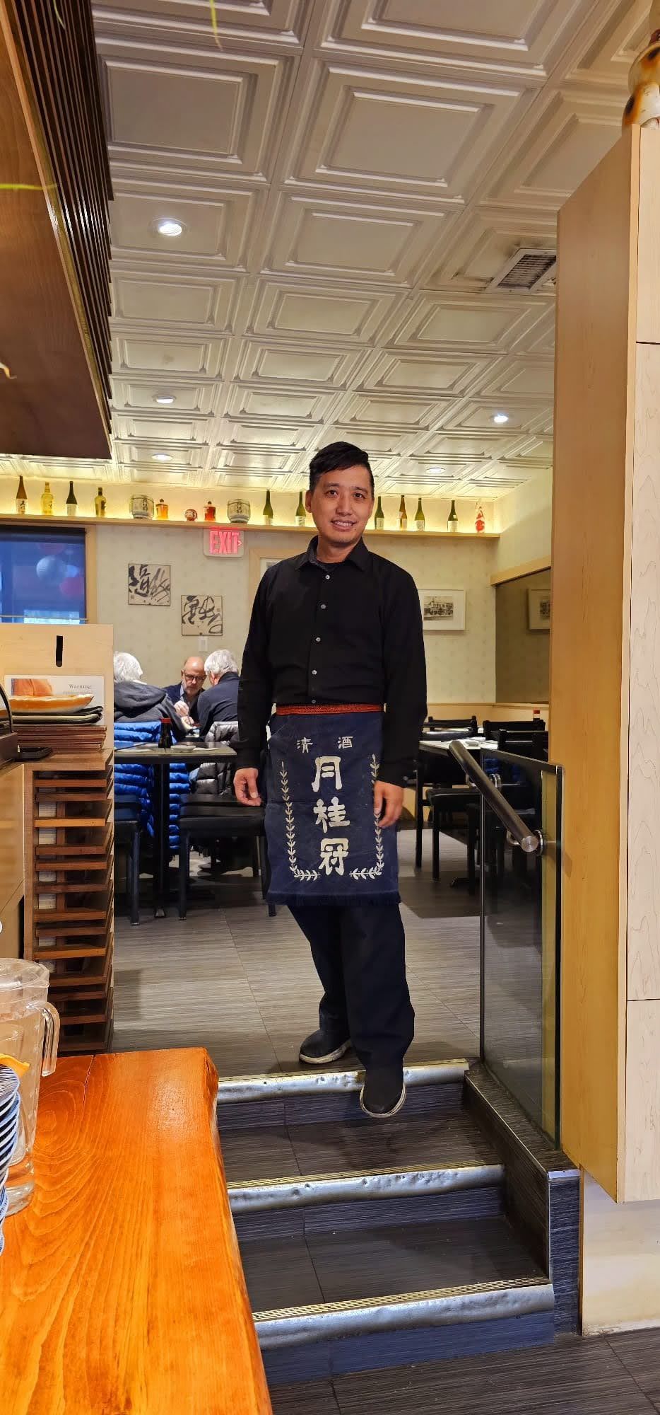 Person standing on stairs inside a restaurant, wearing a black shirt and apron with Chinese characters.