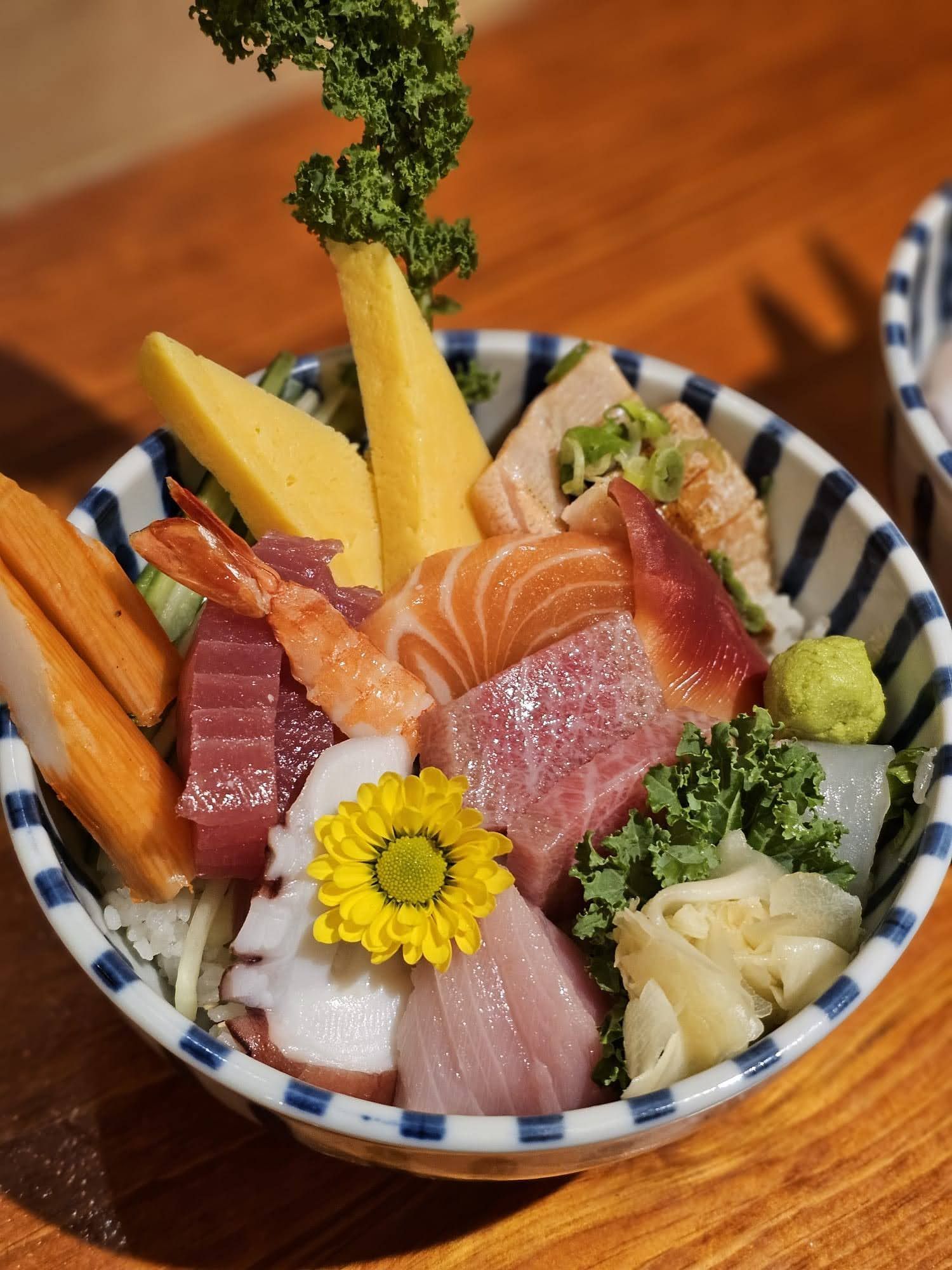 Sushi sashimi bowl with salmon, tuna, shrimp, tamago, wasabi, and a small yellow flower garnish.