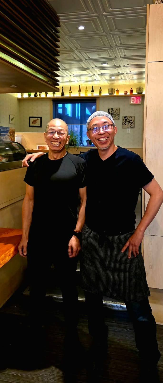 Two people in black shirts smiling in a warmly lit restaurant with a mirrored ceiling