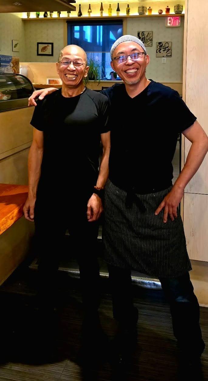 Two people smiling and posing indoors in a warmly lit room, both dressed in black.