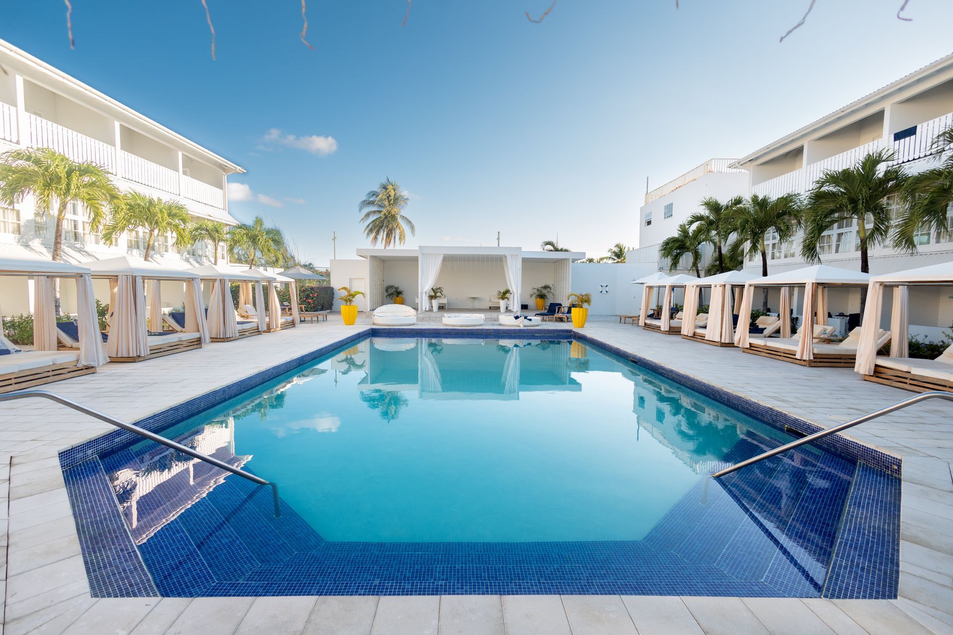 Modern resort courtyard with a blue swimming pool, white buildings, and palm trees under a clear sky
