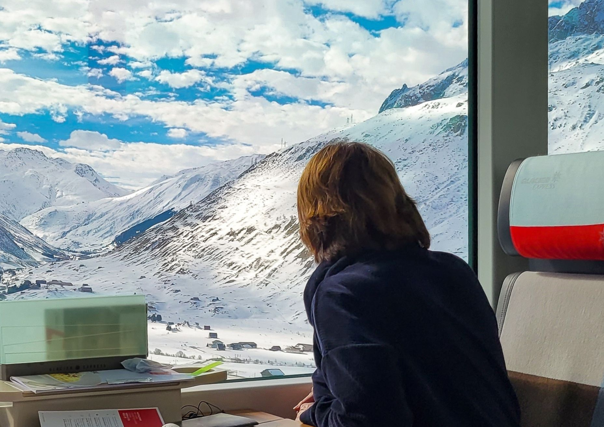 Person gazing out a window at snowy mountains under a blue sky