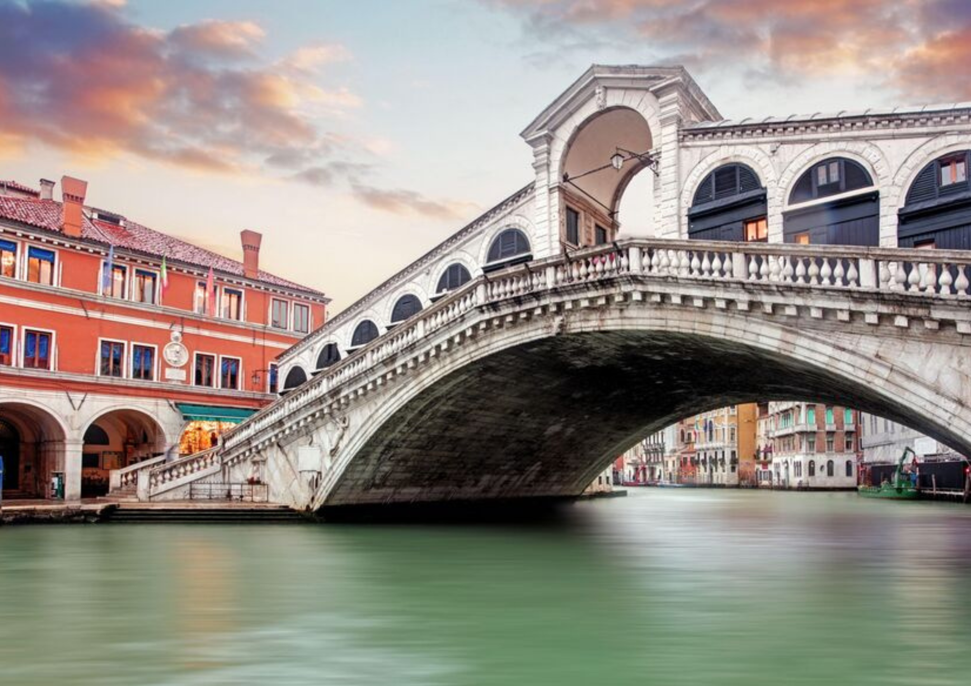 Venetian stone bridge in Venice over a green canal beside red-orange buildings at sunset