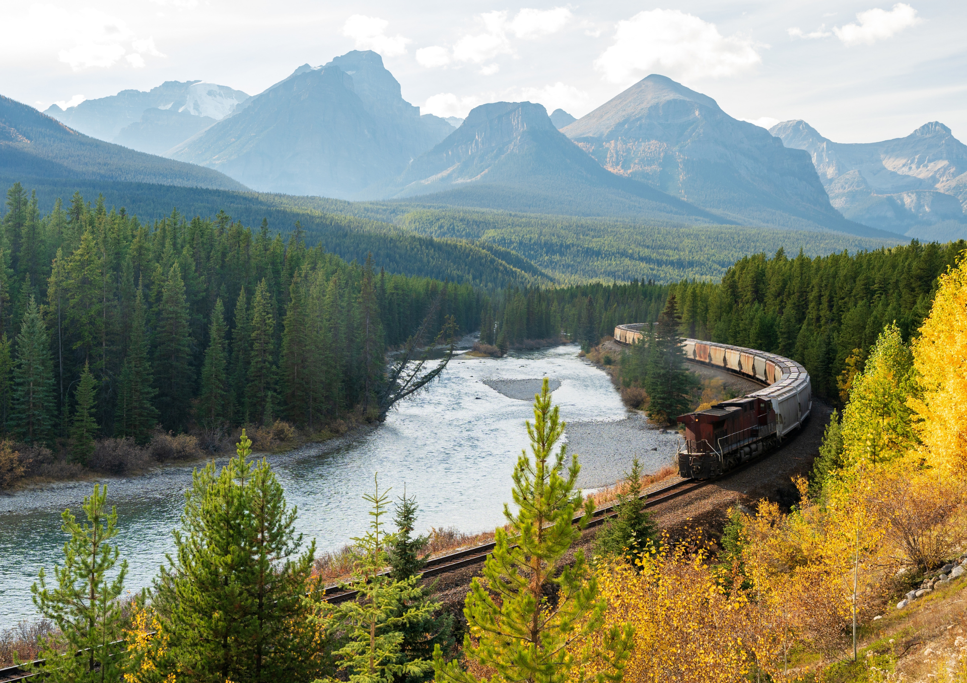 Curving train through a mountain river valley with pine forests and autumn colors