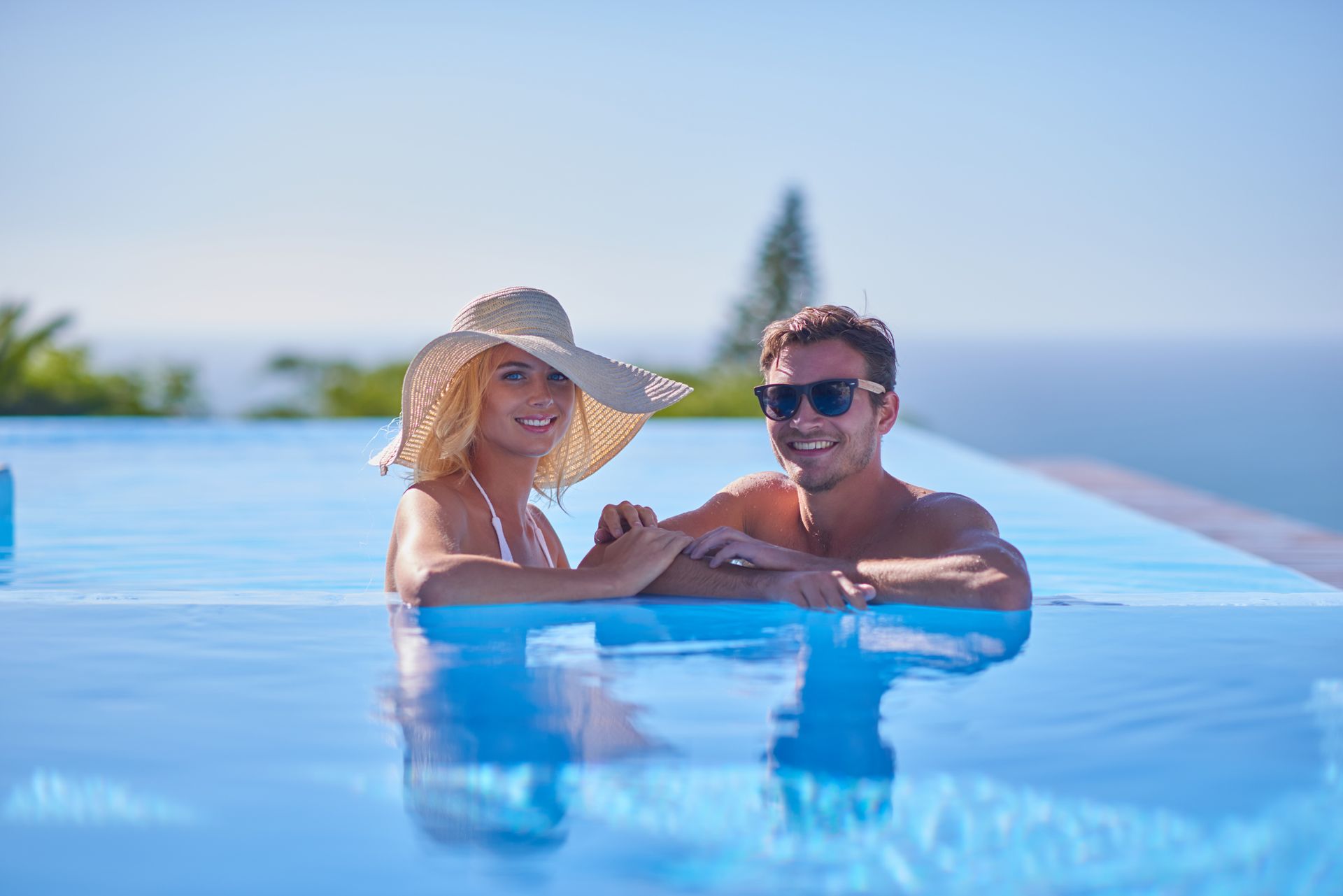 Couple relaxing in an infinity pool under a sun hat, with blue water and a sunny sky.