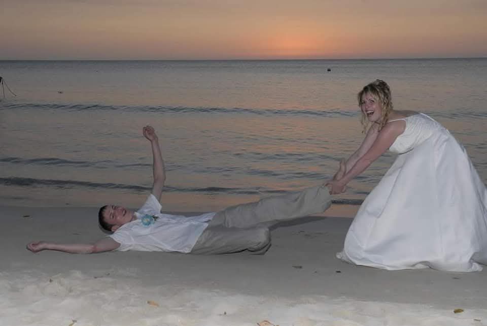 Couple posing on a beach at sunset, one lying in the sand while the other kneels beside them.