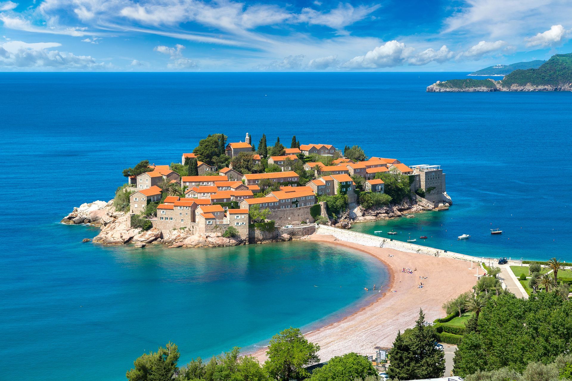 A coastal island town with red-roofed buildings, a sandy causeway, and turquoise sea under a blue sky