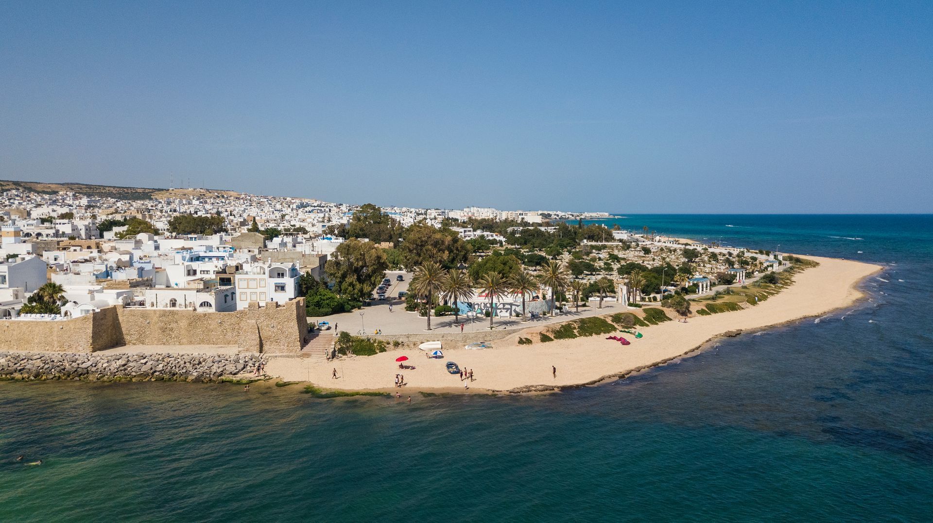 Coastal town with white buildings, sandy beach, and turquoise sea along a curved shoreline