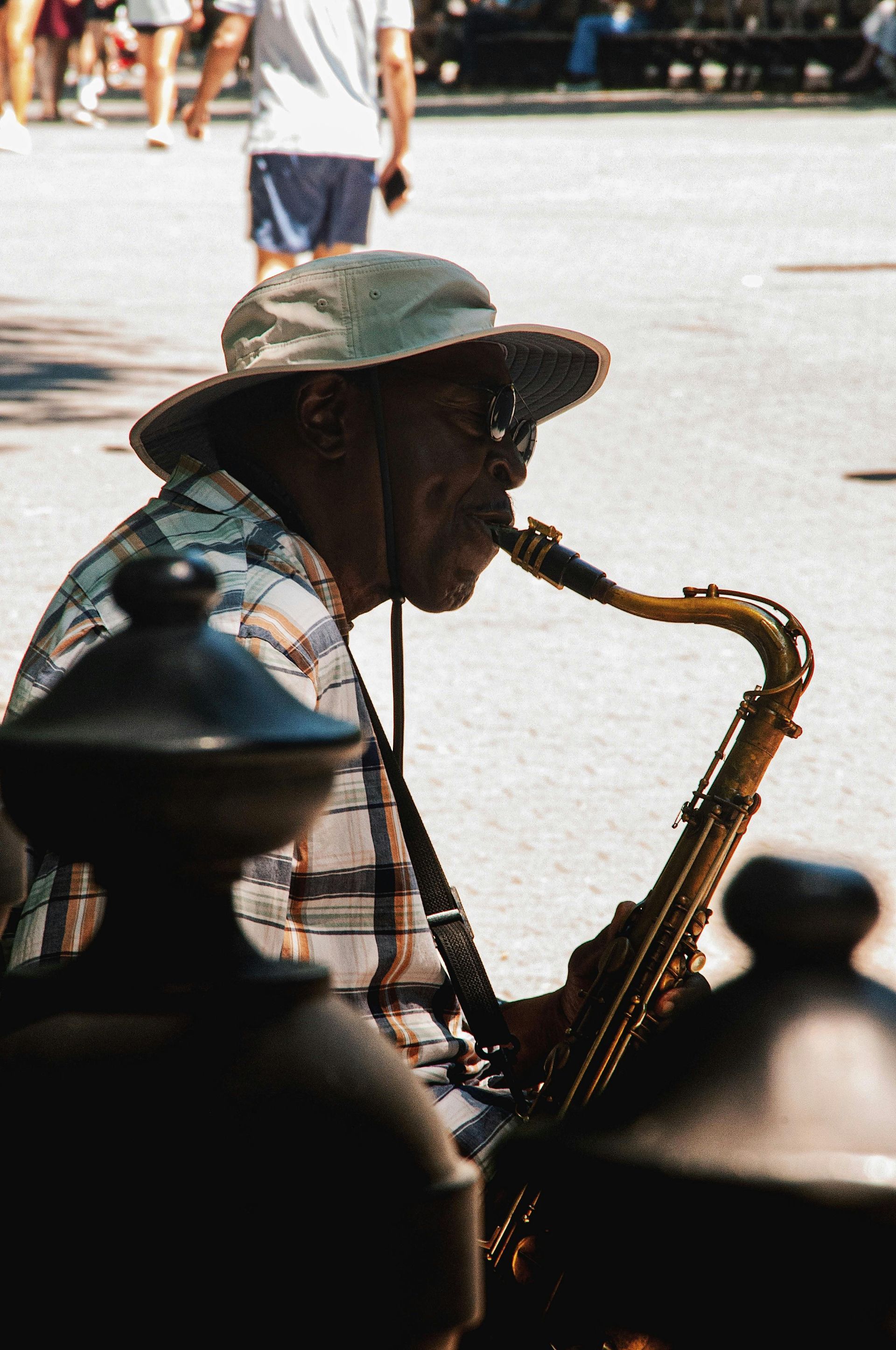 Street saxophonist in a hat playing on a sunny sidewalk, with blurred pedestrians in the background.
