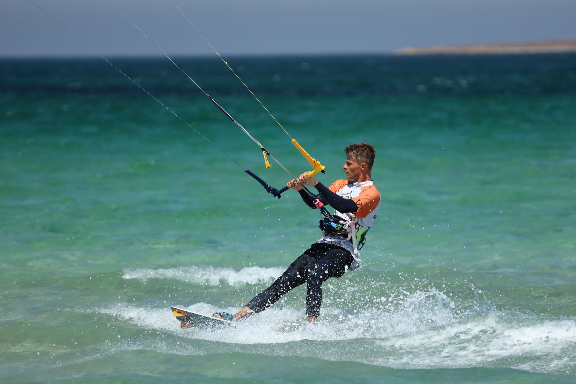 Kite surfer carving across turquoise waves, spray splashing against a sunny beach horizon