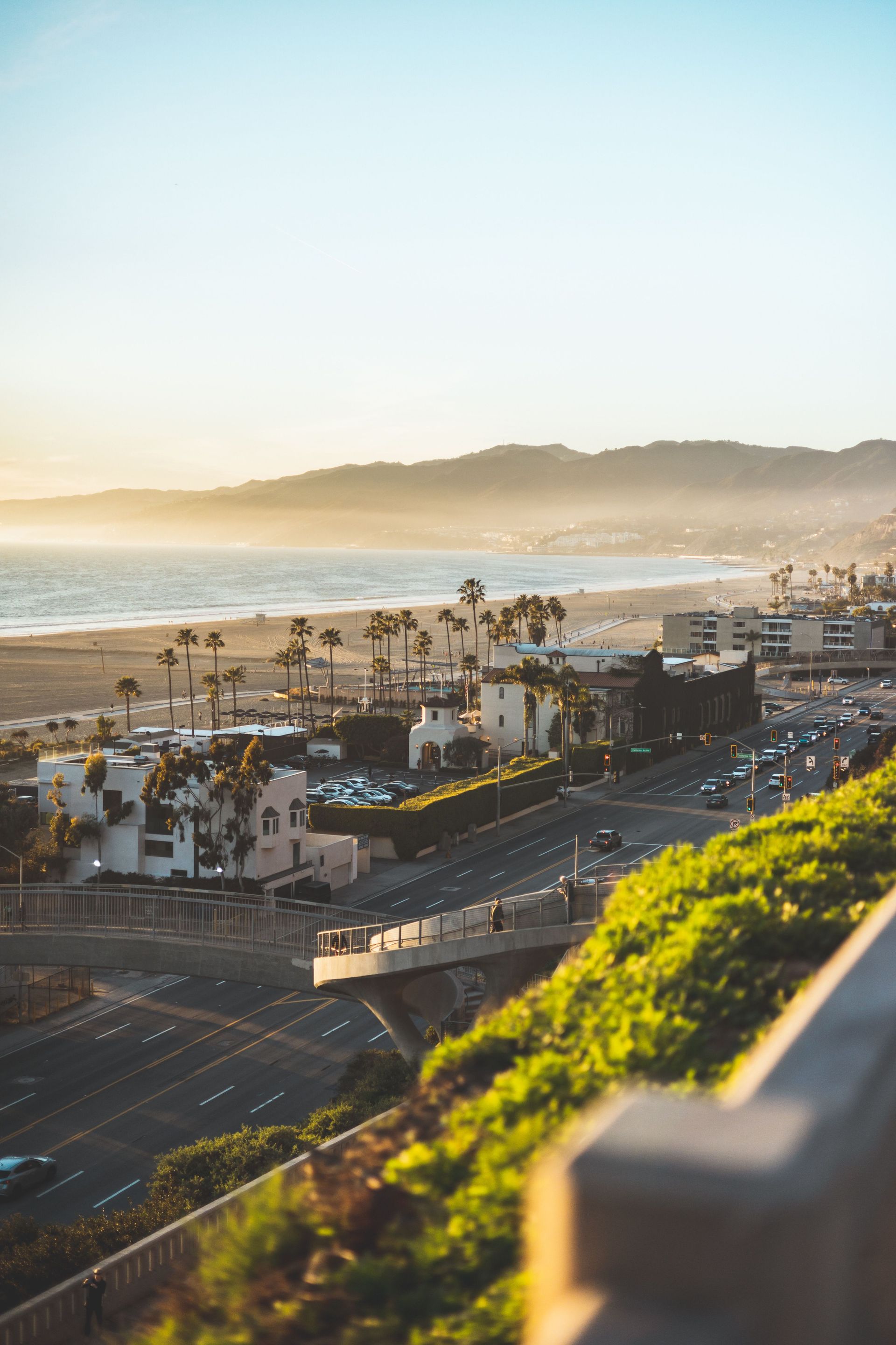 Sunset over a beach promenade with palm trees, parked cars, and ocean waves in the distance