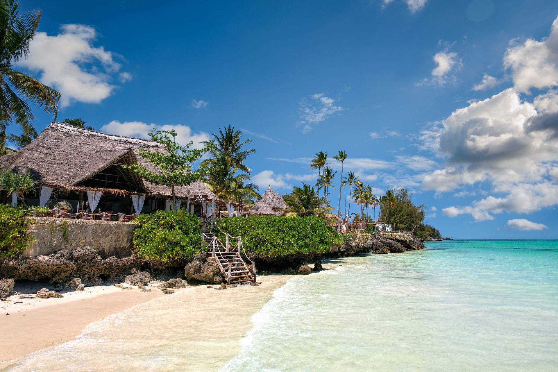 Tropical beach with white sand, turquoise water, palm trees, and a thatched-roof resort under a blue sky