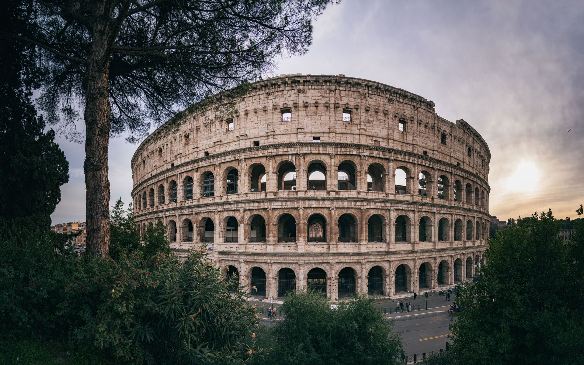 The Colosseum in Rome at sunset, framed by trees and a cloudy sky.