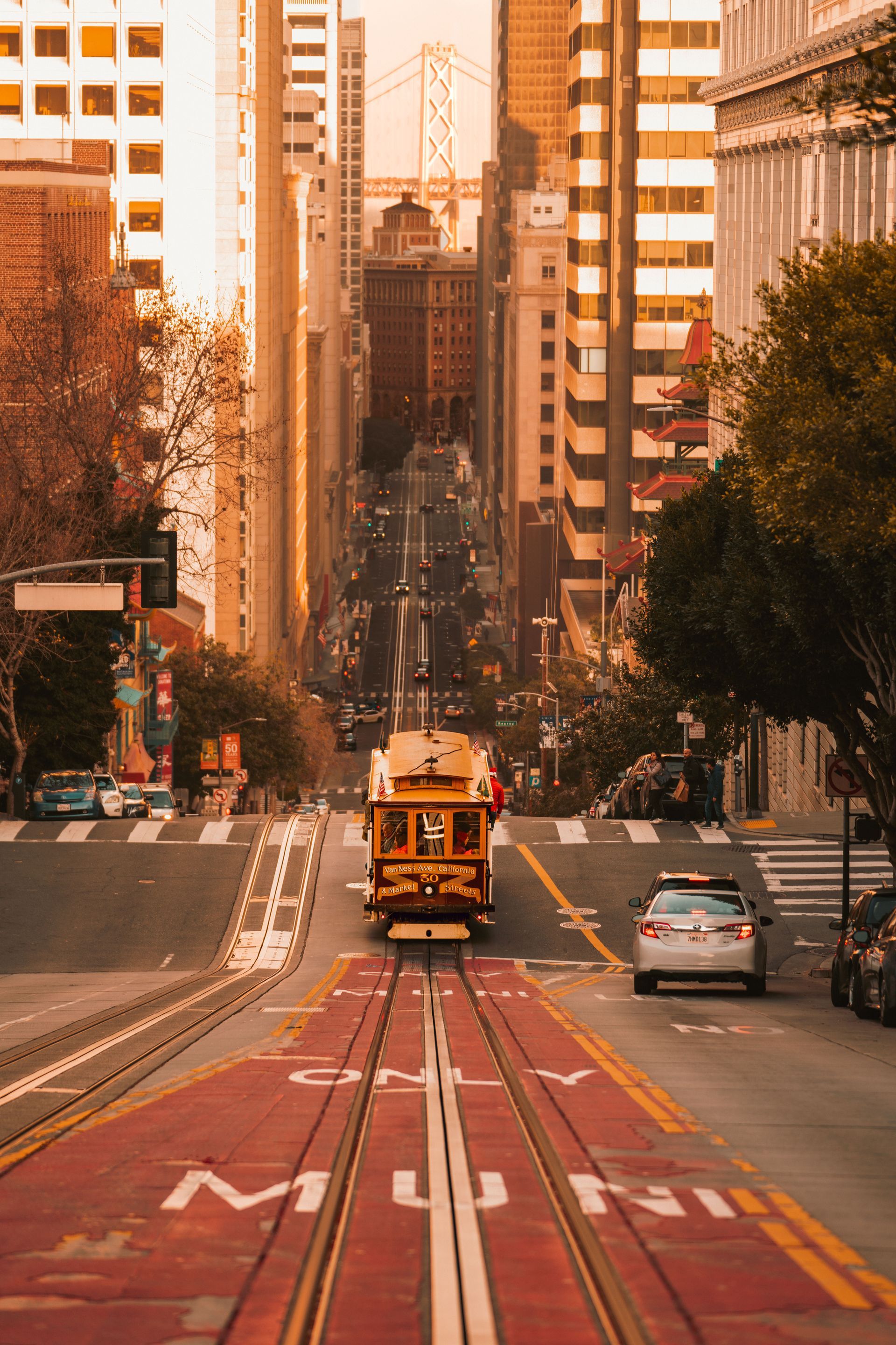 Cable car descending a steep city street between tall buildings, with cars and red transit lanes.