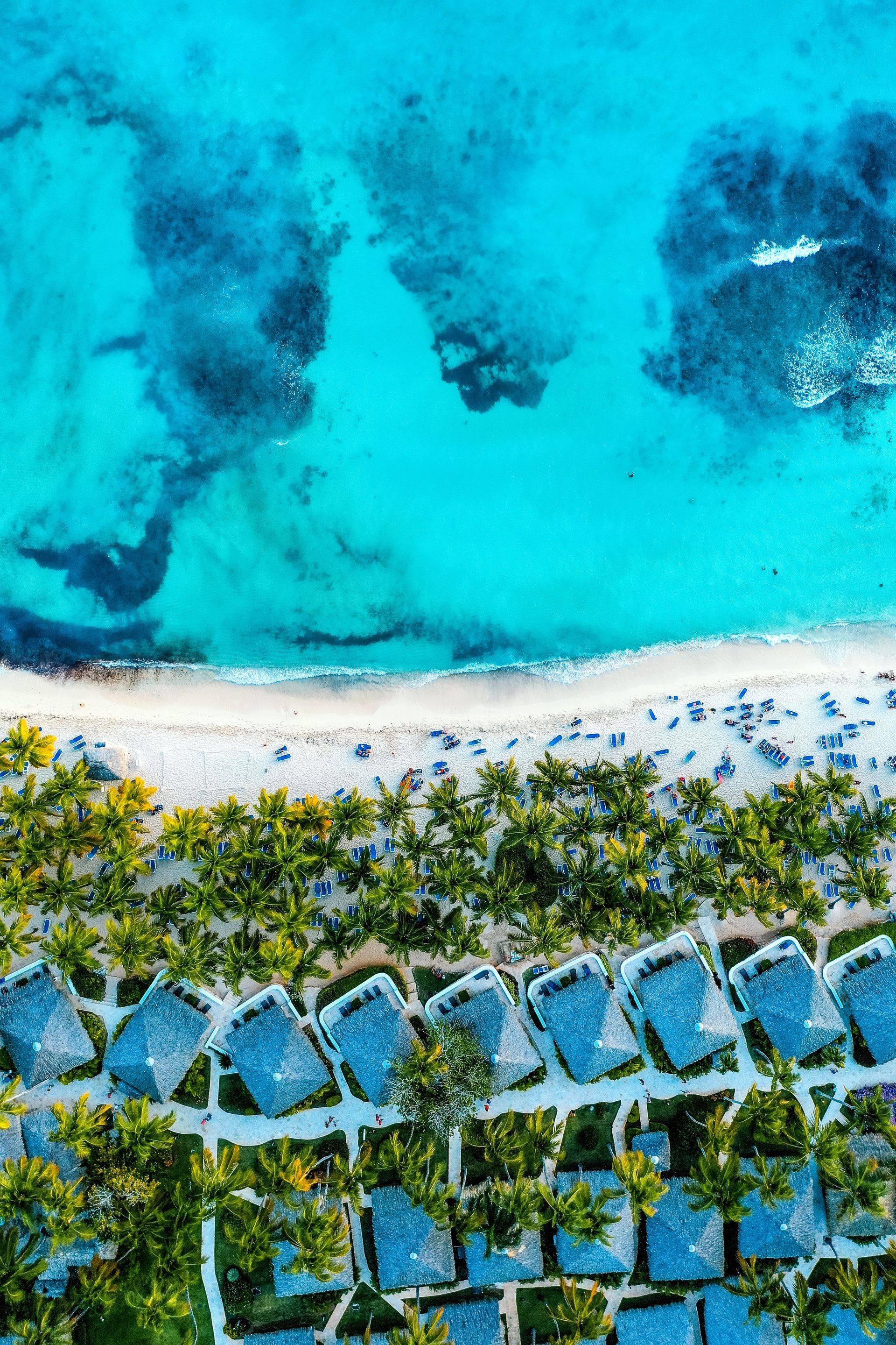 Aerial view of turquoise ocean, white sandy beach, and palm trees with blue-roofed huts.