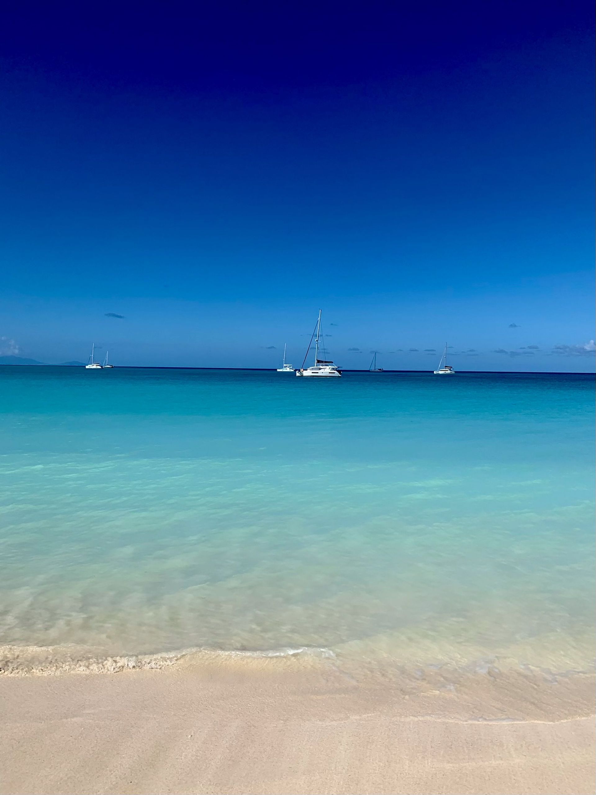 Calm tropical beach with turquoise water, white sand, and sailboats on the horizon under a deep blue sky