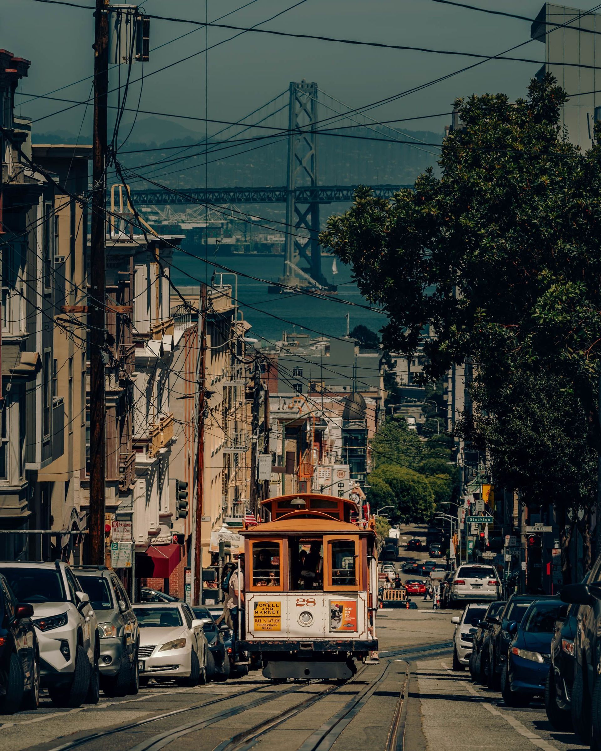 Orange cable car on a steep city street with parked cars, overhead wires, and a bridge in the distance