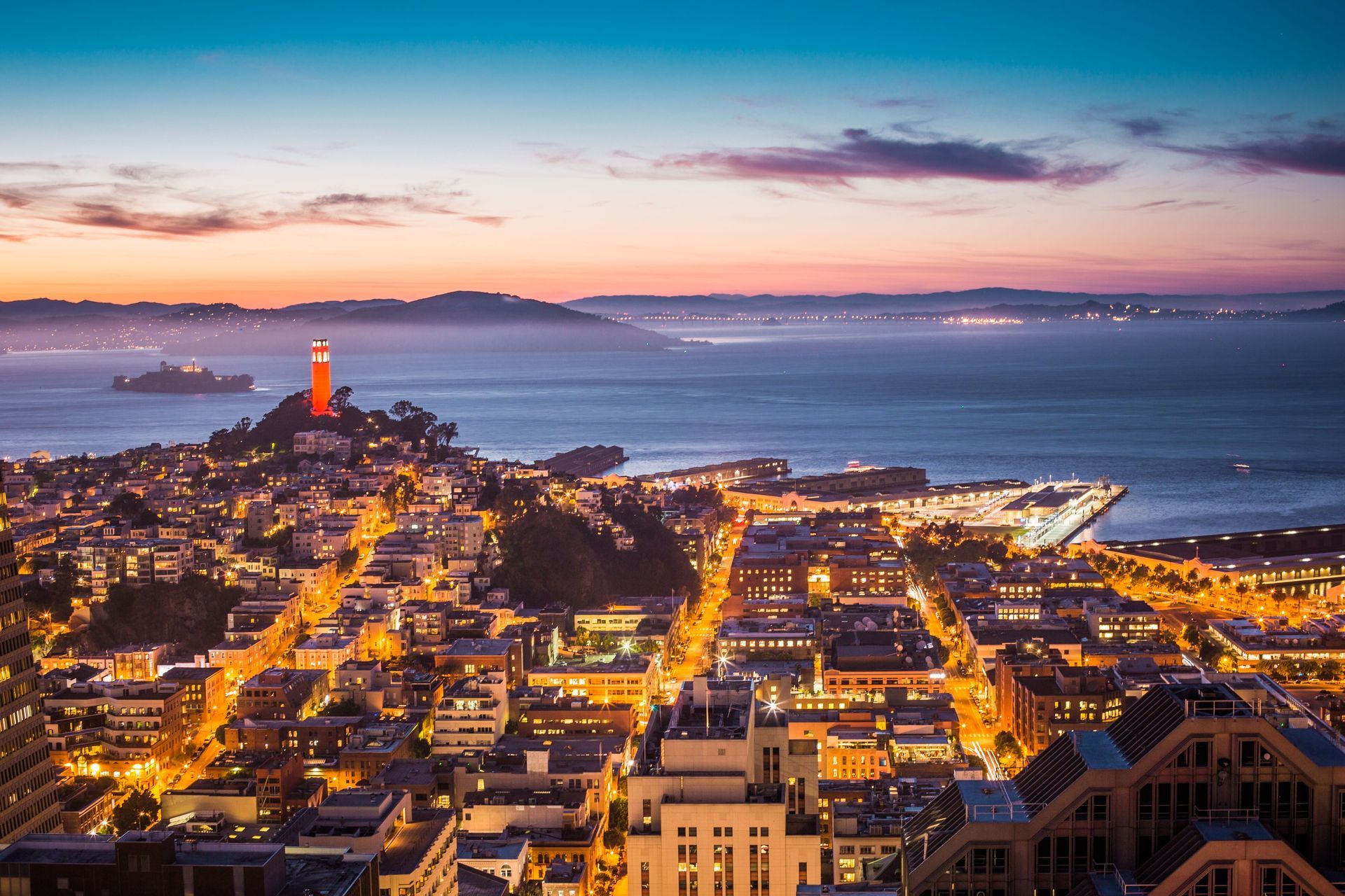 Sunset over a coastal city harbor with glowing streets, a lighthouse, and calm blue water.