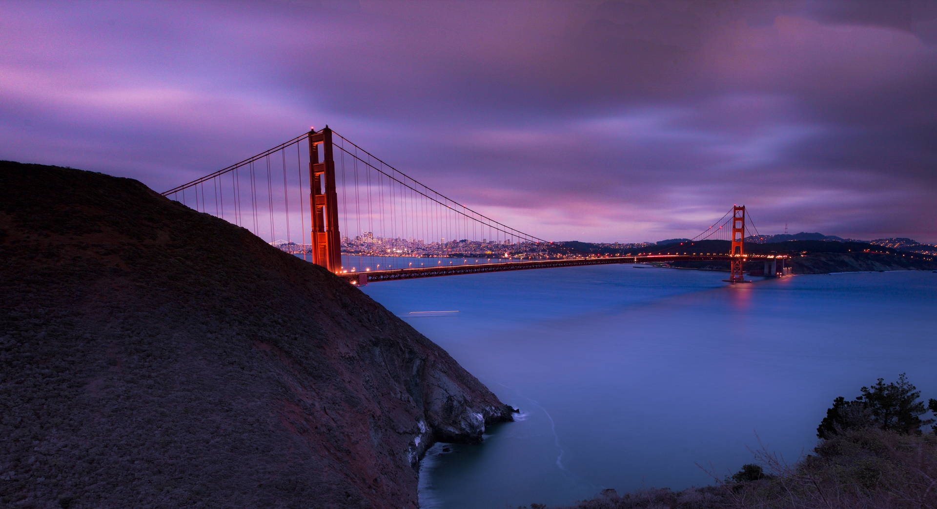 Golden Gate Bridge at dusk, glowing orange against a purple sky over calm blue water.