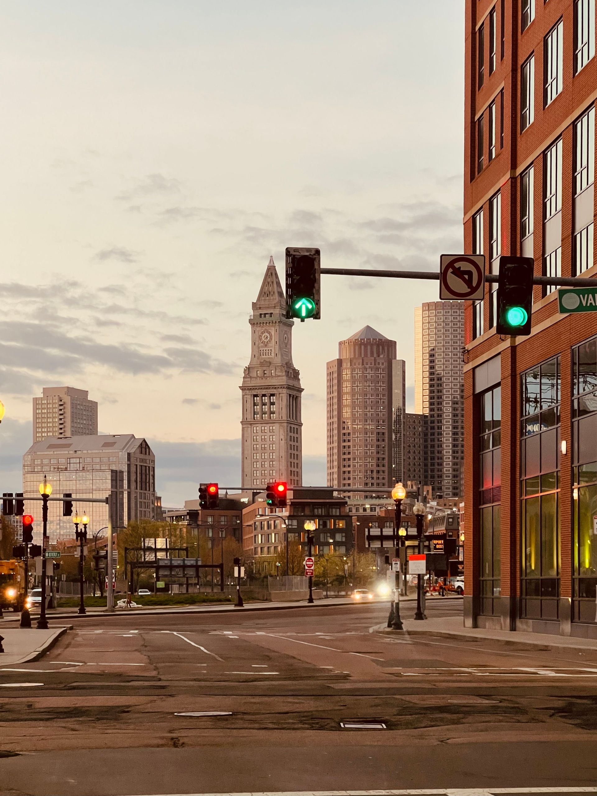 Downtown city street at dusk with traffic lights, tall buildings, and a clock tower in the distance